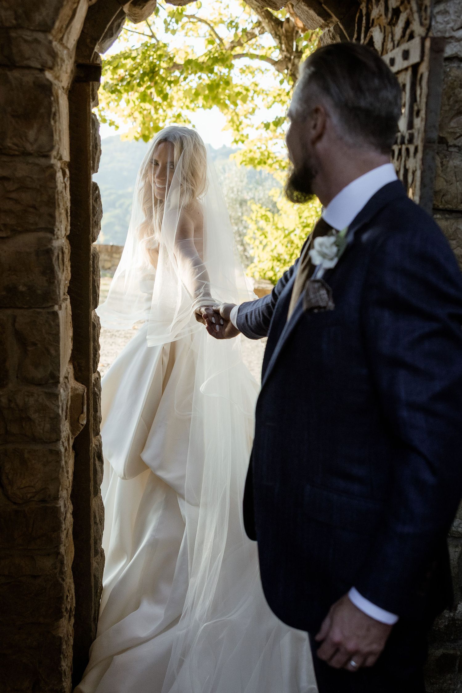 A bride in a white wedding dress and veil stands in an arched stone passageway with sunlight streaming through.