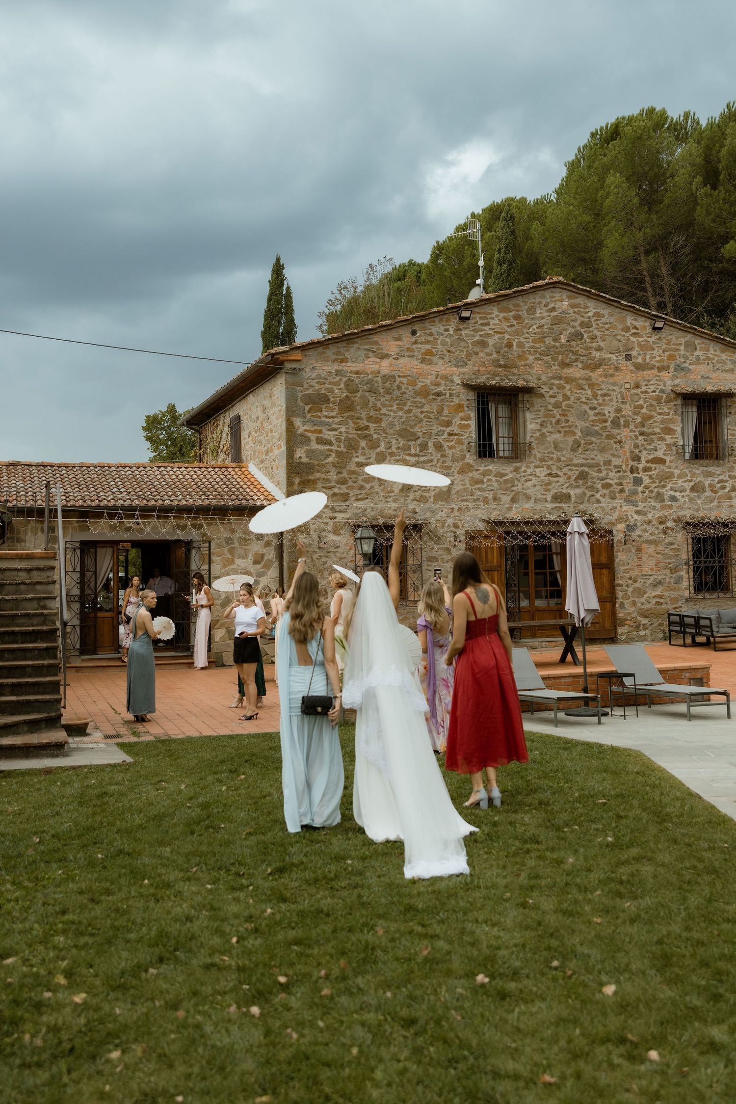 Wedding party gathers outside rustic stone farmhouse under cloudy skies on grassy lawn.