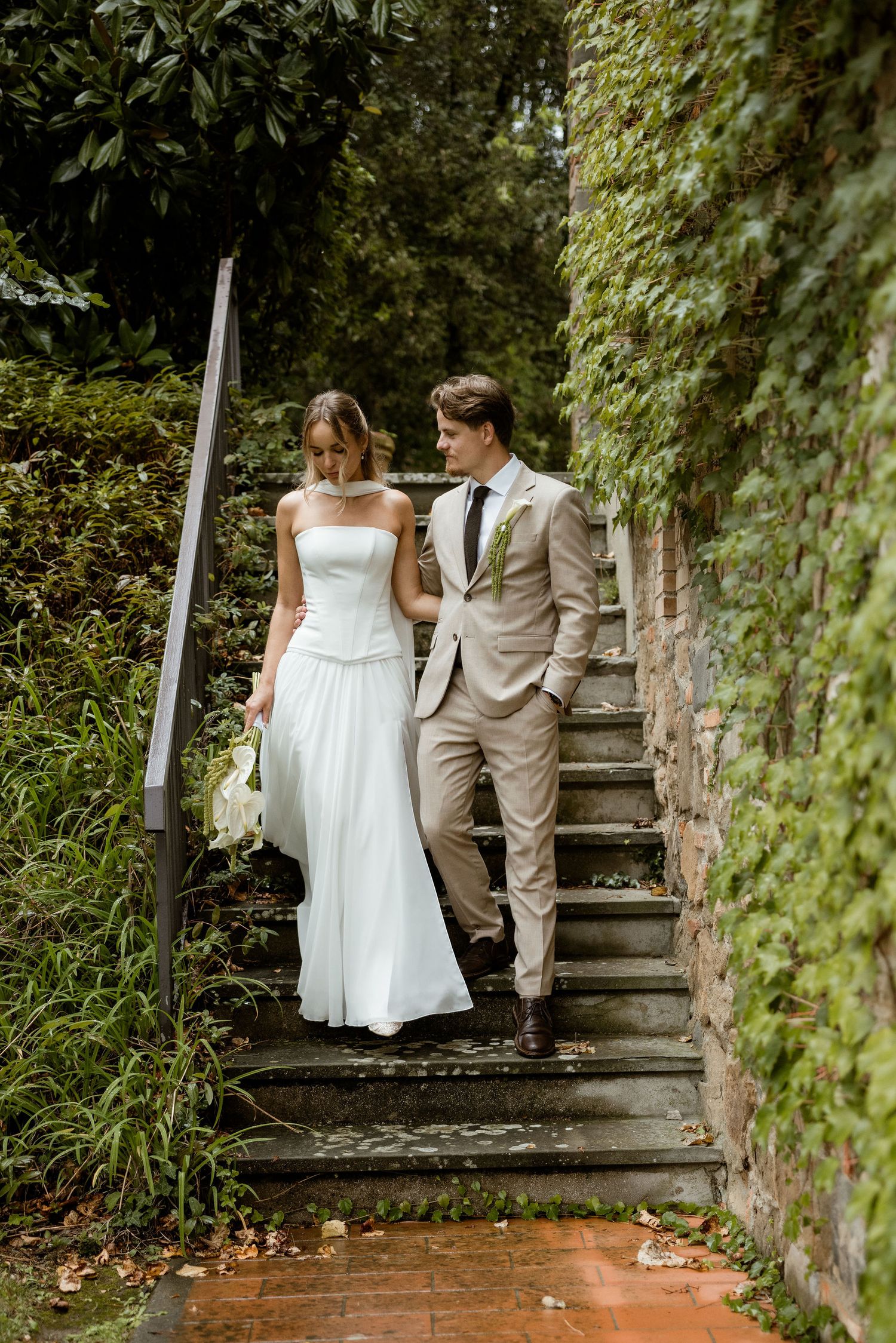 A couple descends stone garden steps surrounded by lush greenery during a wedding photoshoot.