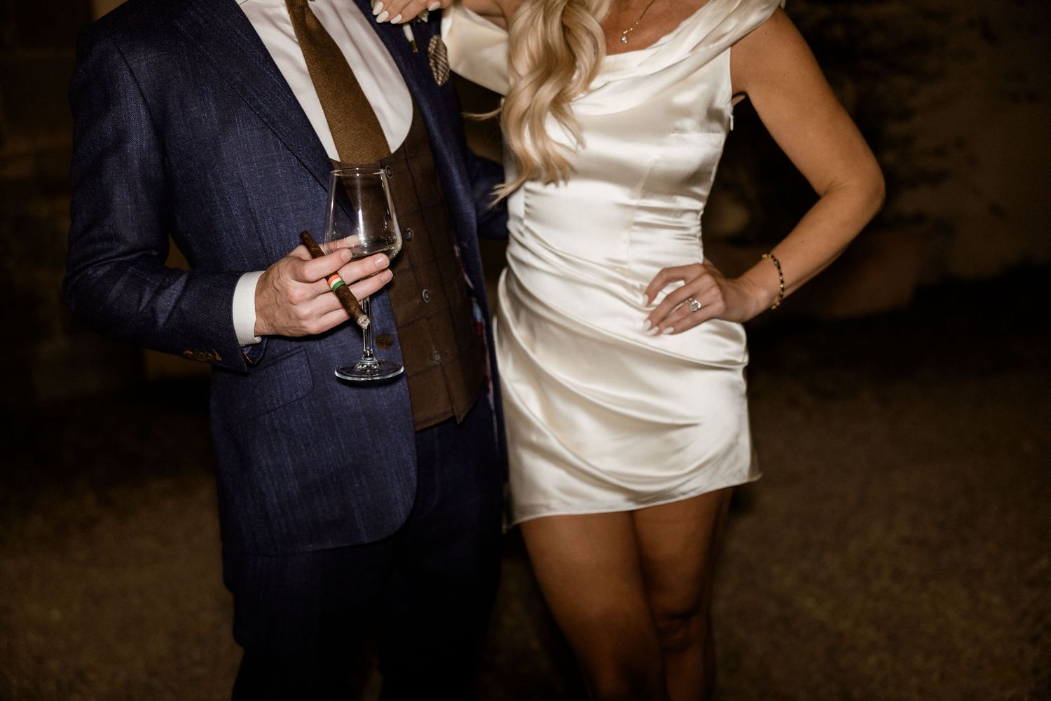 Couple in formal attire holding wine glasses at an evening event, with navy suit and short white satin dress.