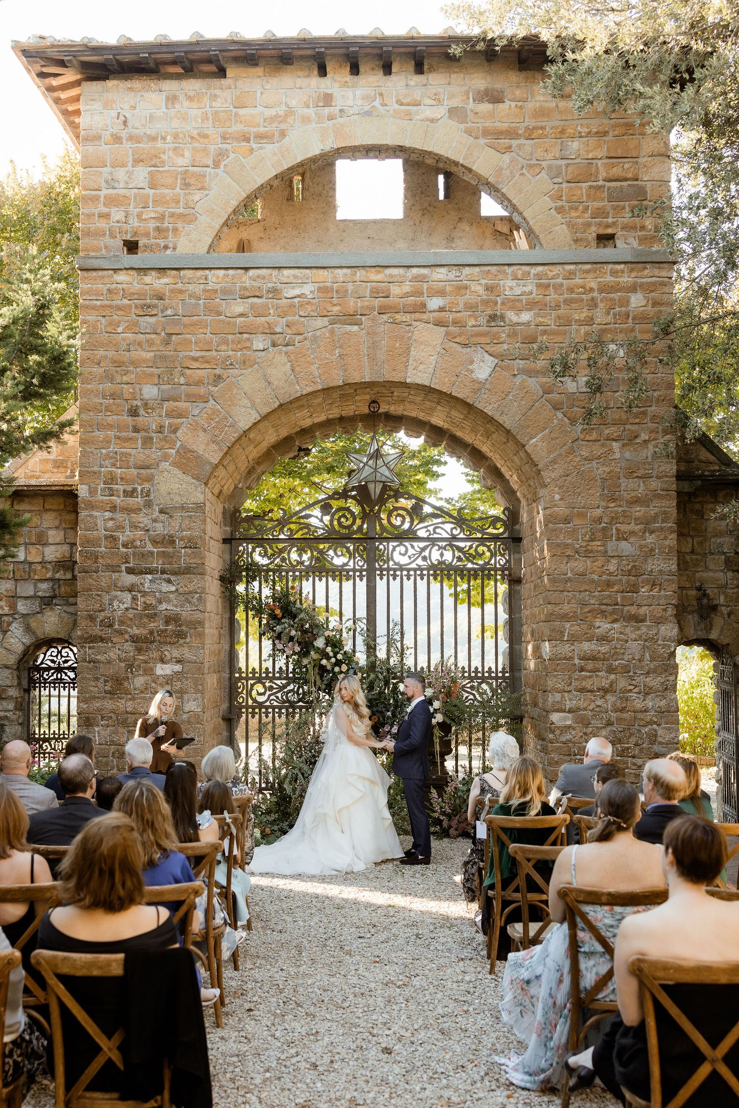 Outdoor wedding ceremony at rustic stone venue with arched gateway and wooden chairs set up on gravel path.