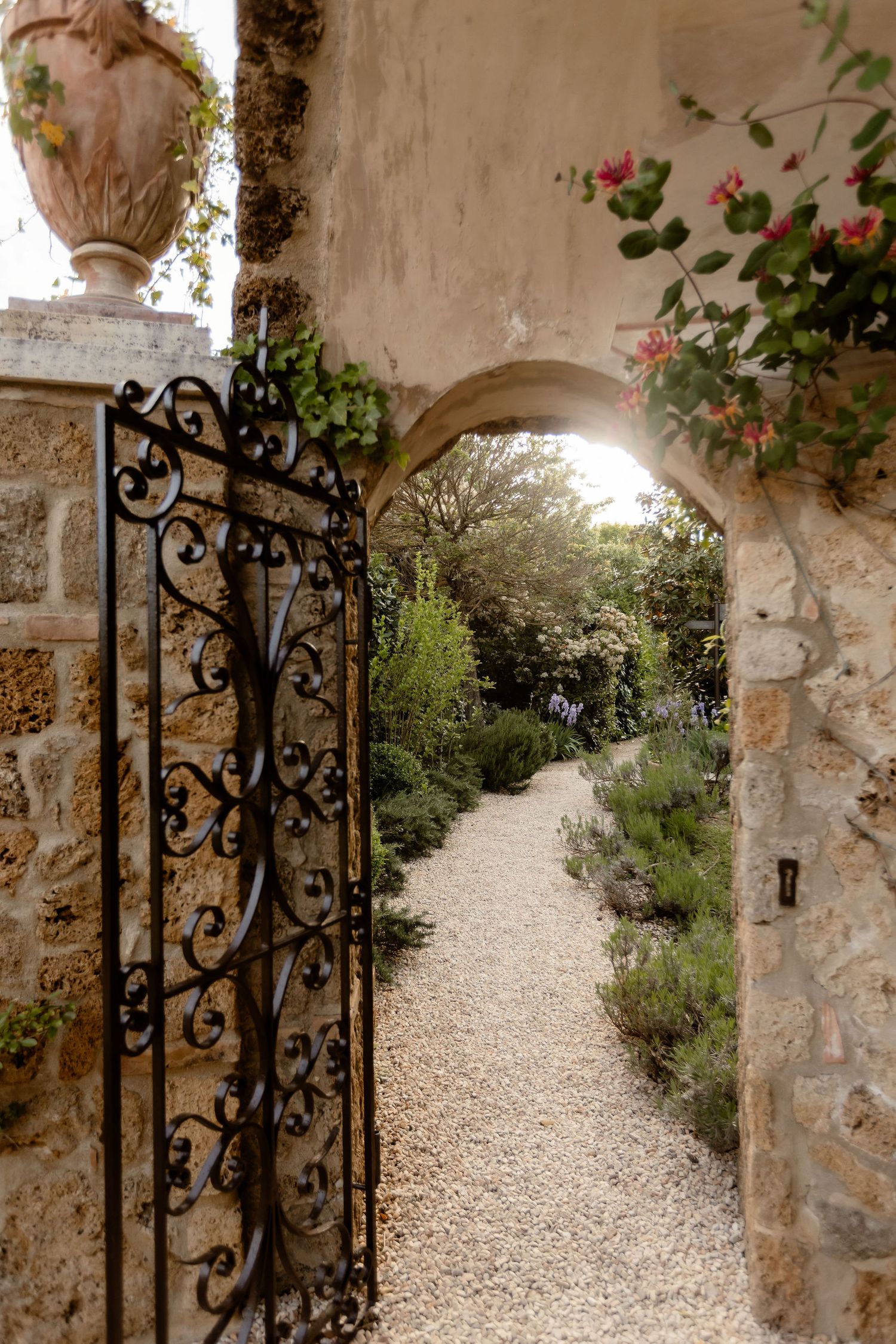 Ornate wrought iron garden gate opens to a gravel path through an arched stone entrance with climbing roses and greenery.