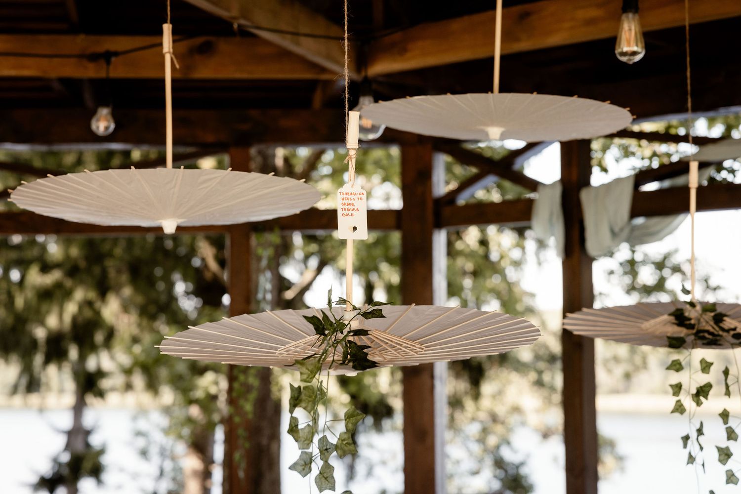 Modern rustic light fixtures with white umbrella-shaped shades hang from wooden beams in an outdoor setting.