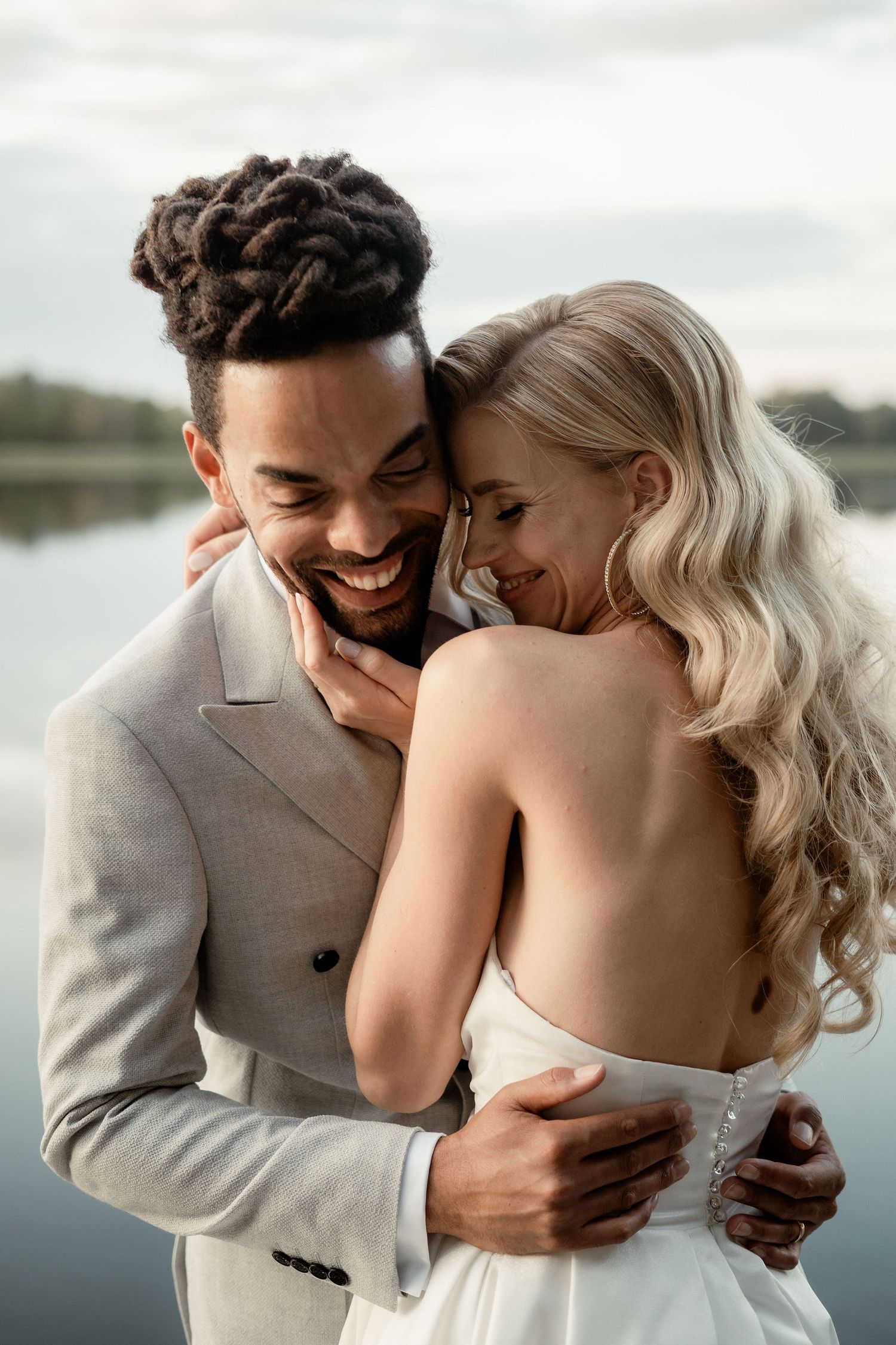 A happy couple shares an intimate moment by a lake, with the person in a gray suit embracing their partner in white.