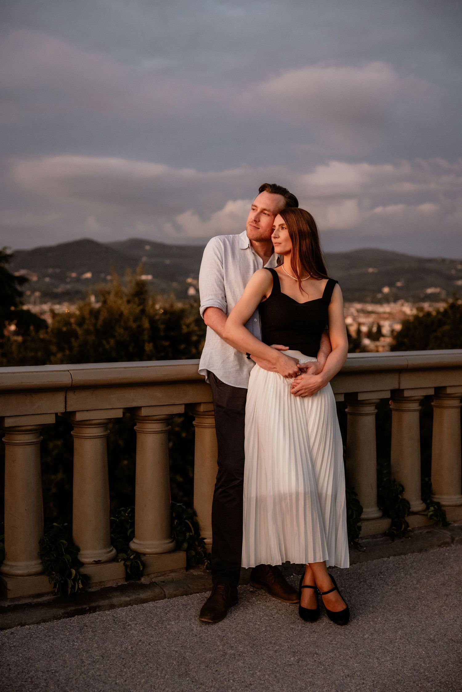 A couple embraces while posing against a stone balustrade during golden hour with a city vista behind them.