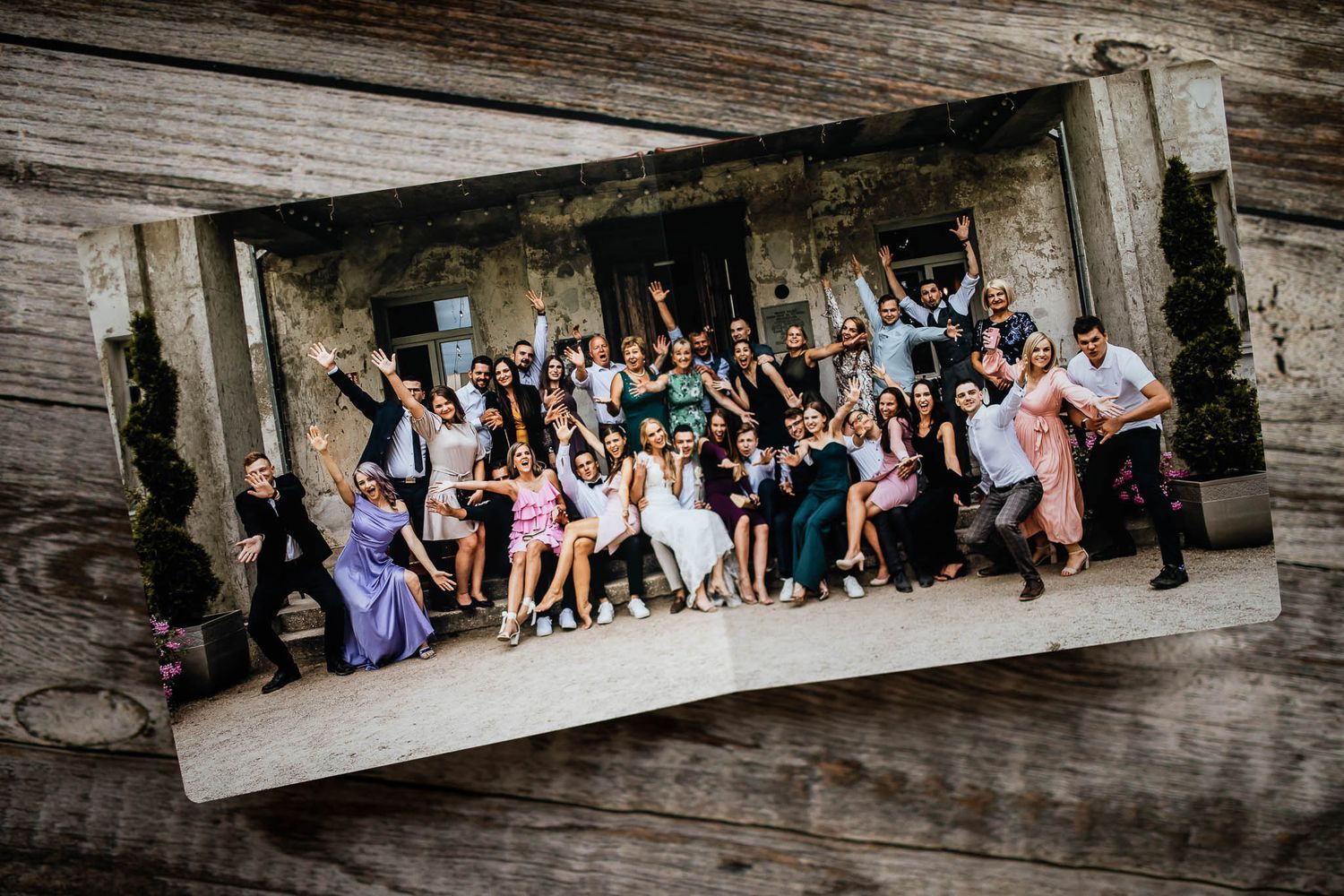 Large wedding party poses together in front of rustic stone building with everyone raising their arms in celebration.