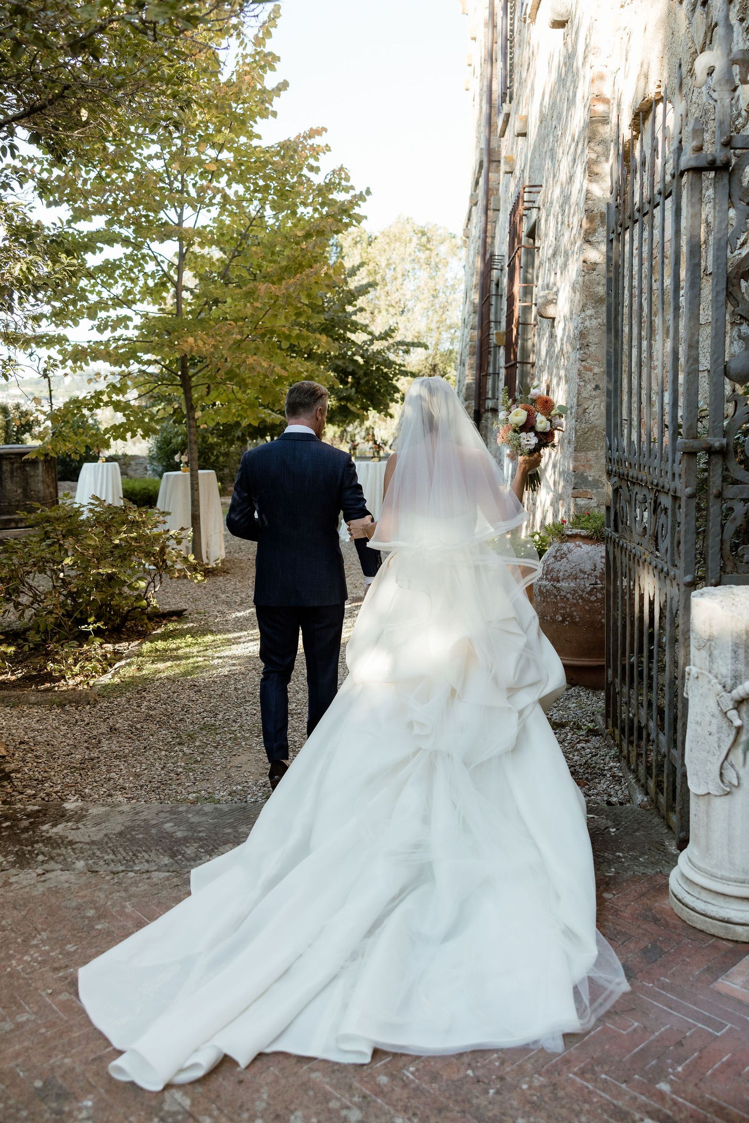 A couple in wedding attire walk along a stone path beside an ornate church exterior with autumn trees in the background.