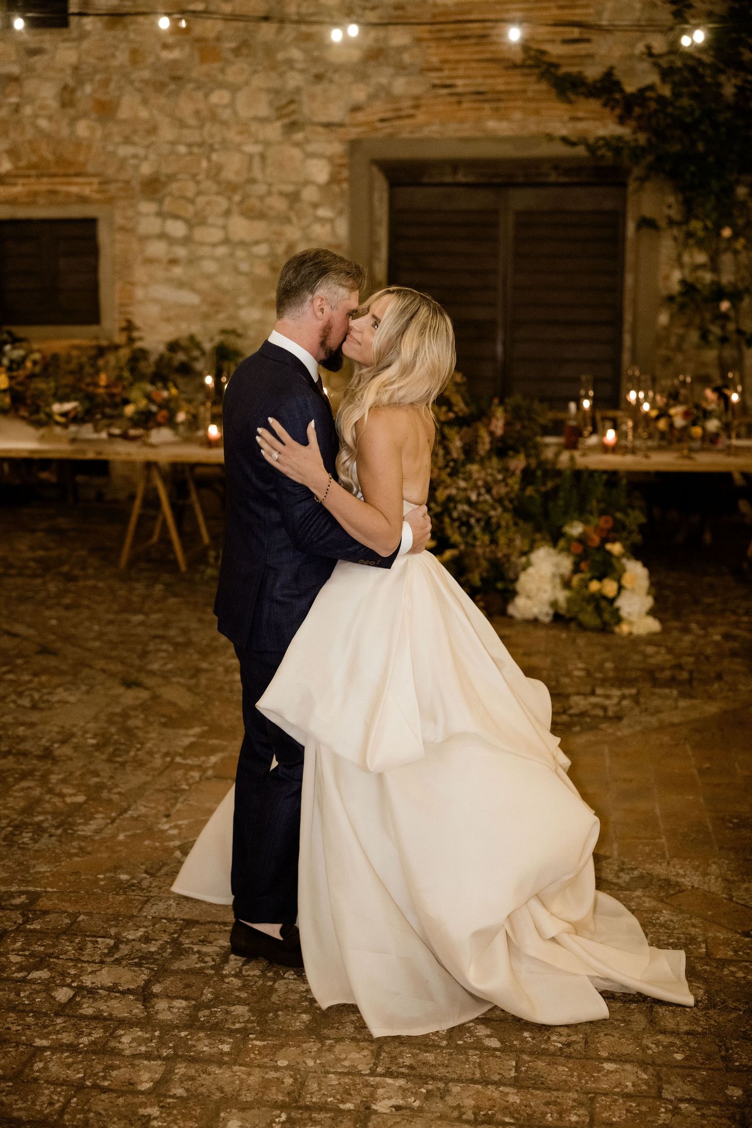 A romantic wedding couple shares an intimate kiss on a rustic stone floor with warm lighting and floral decor.