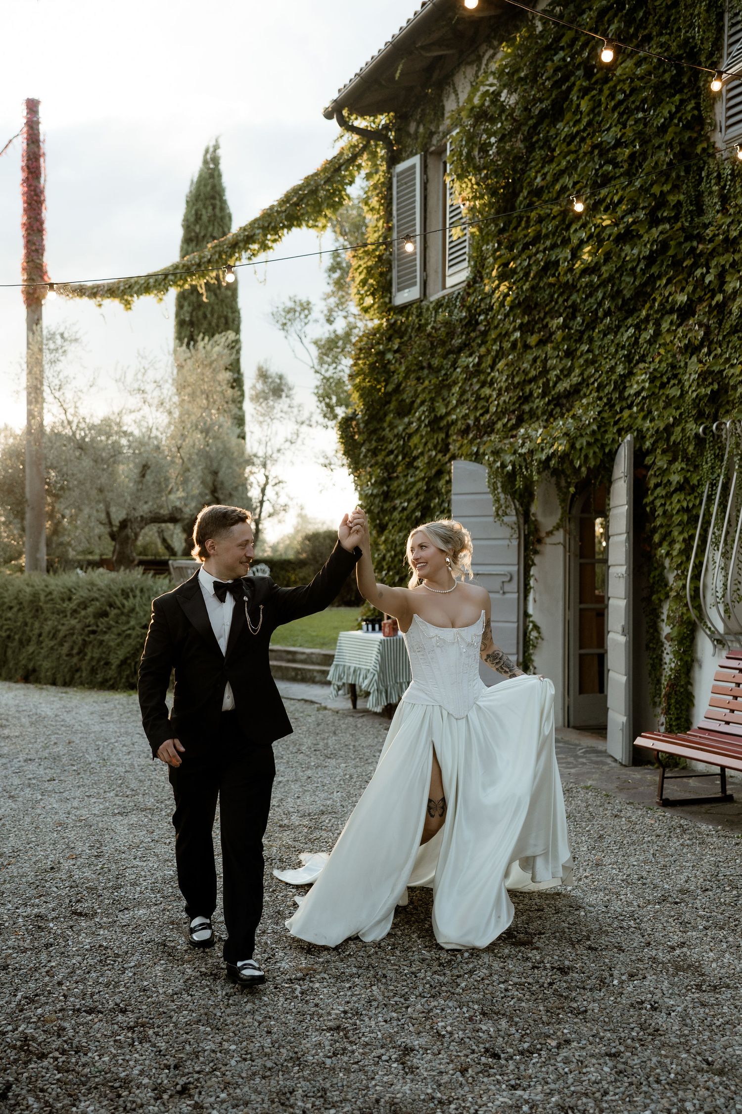 A couple in formal wedding attire walks together through a gravel courtyard surrounded by ivy-covered walls at sunset.