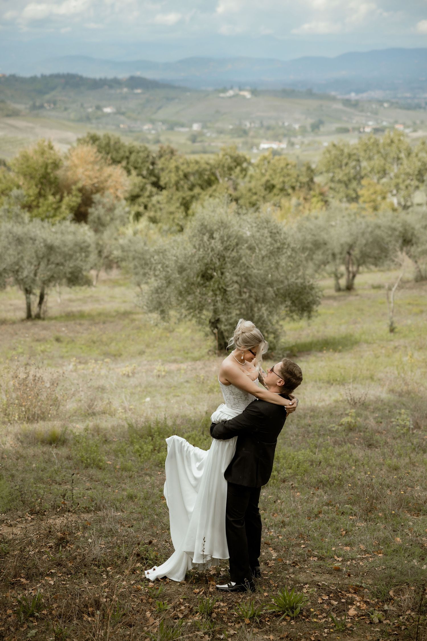 A couple embraces in a romantic dance pose against a scenic backdrop of rolling hills and olive trees in the countryside.