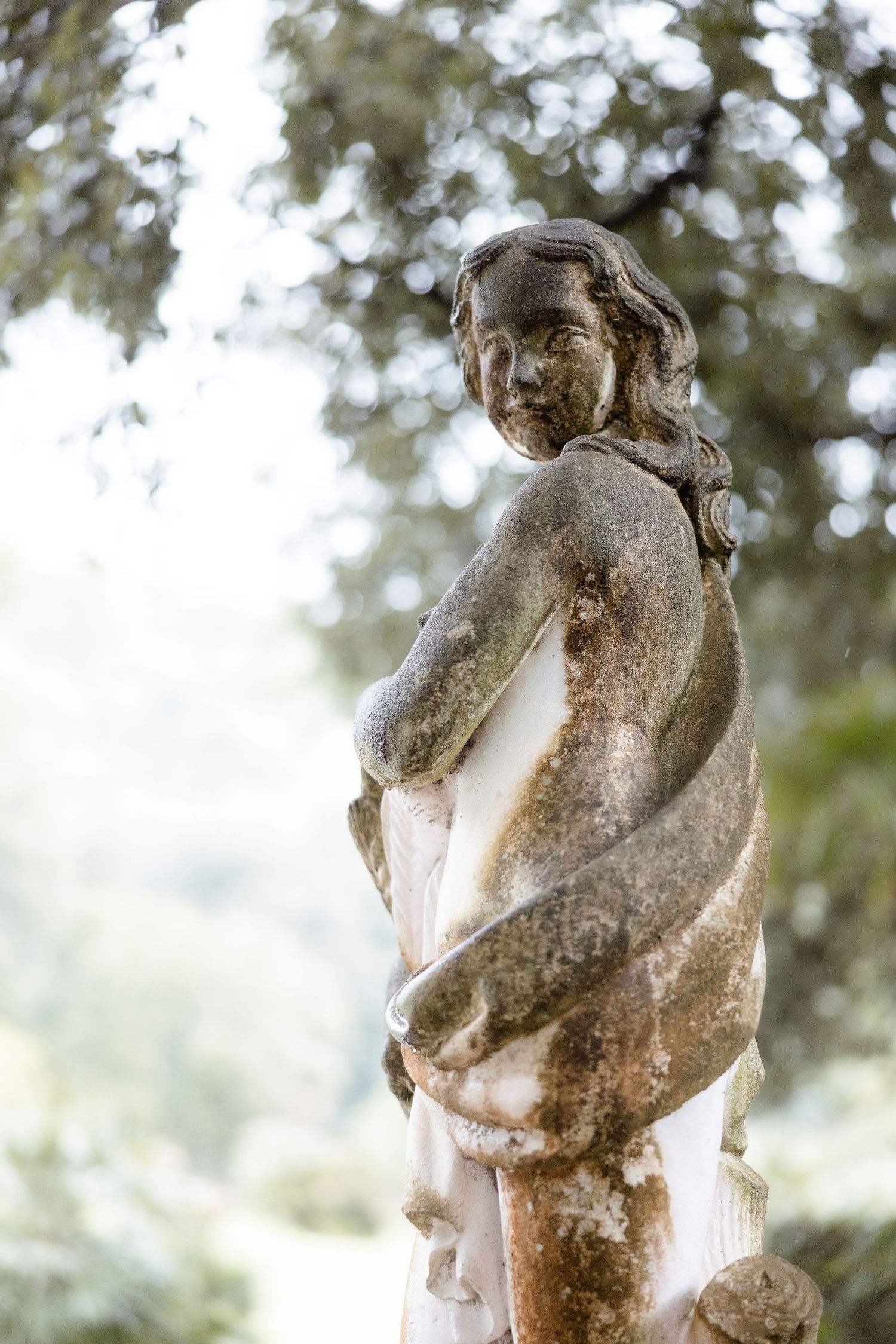 A weathered stone garden statue with a flowing dress stands amid blurred foliage in soft natural lighting.