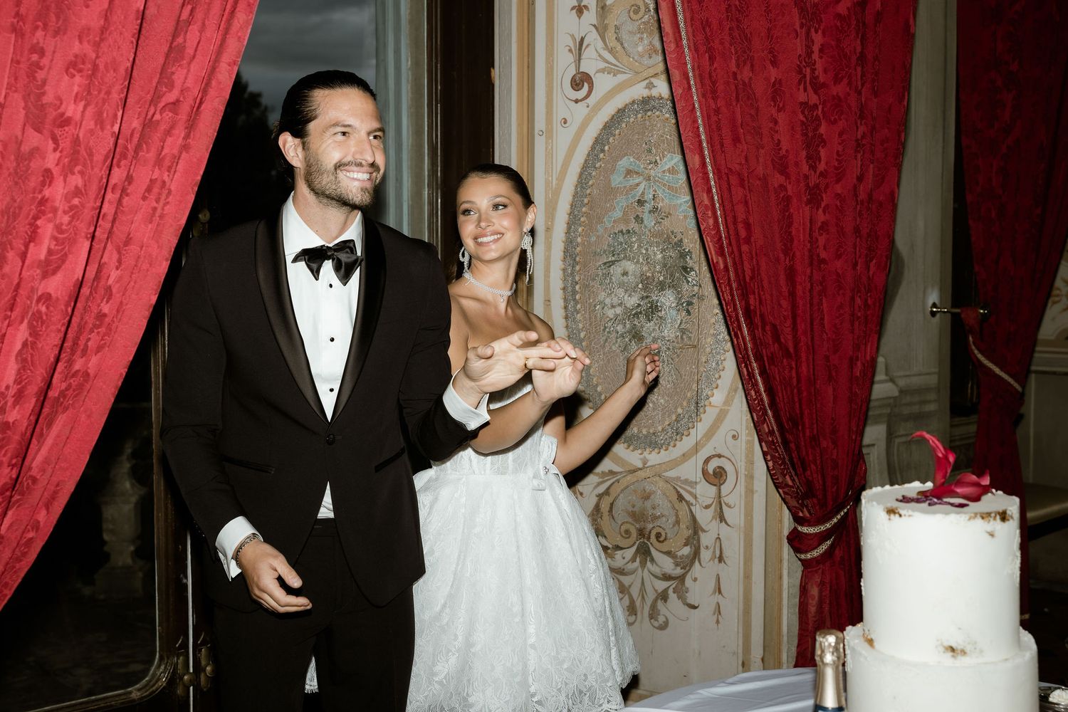 Elegant wedding couple stands under red velvet curtains in an ornate ballroom with gilded architectural details.
