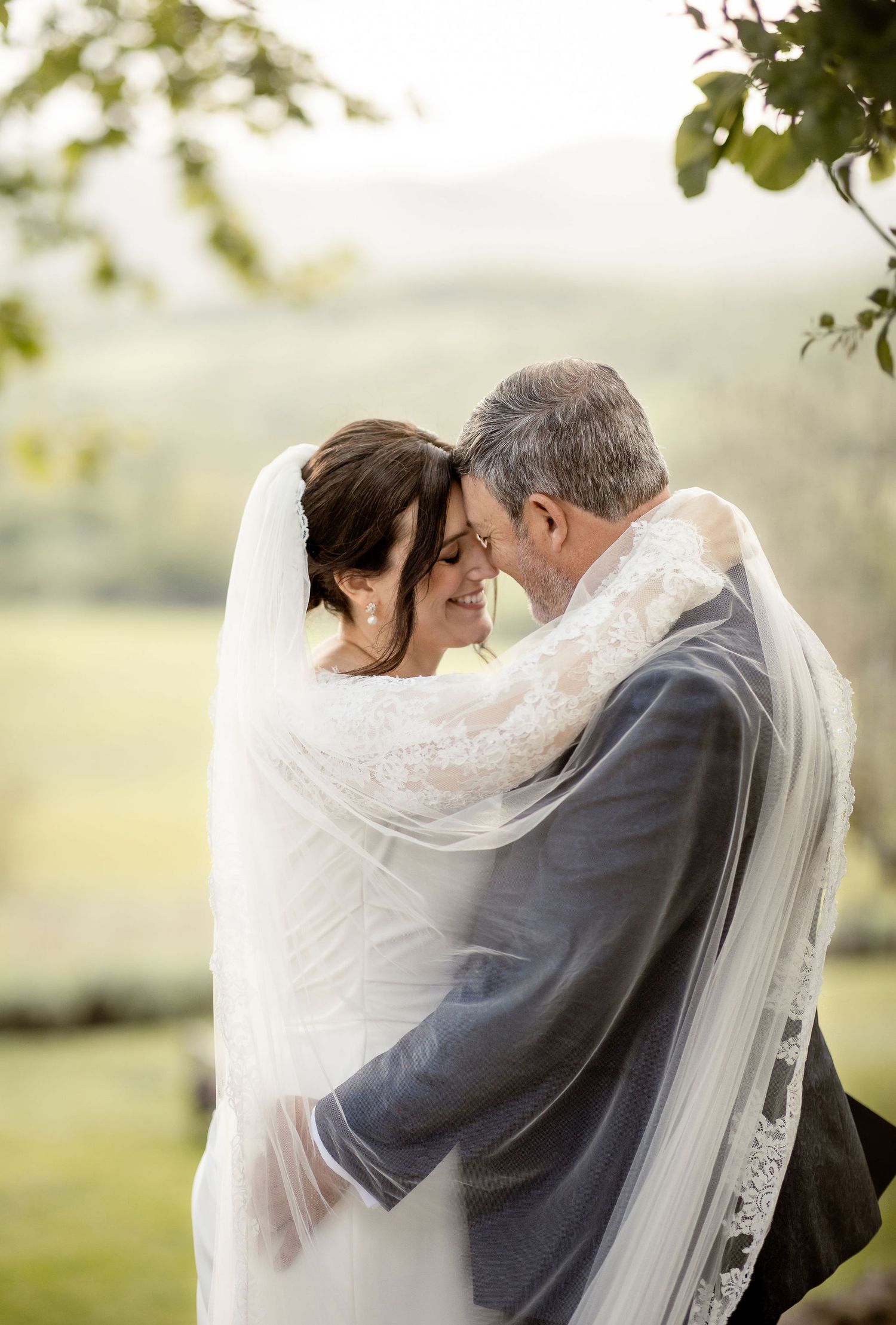 A tender wedding moment captured outdoors with a couple embracing under a flowing white veil against a soft natural backdrop.