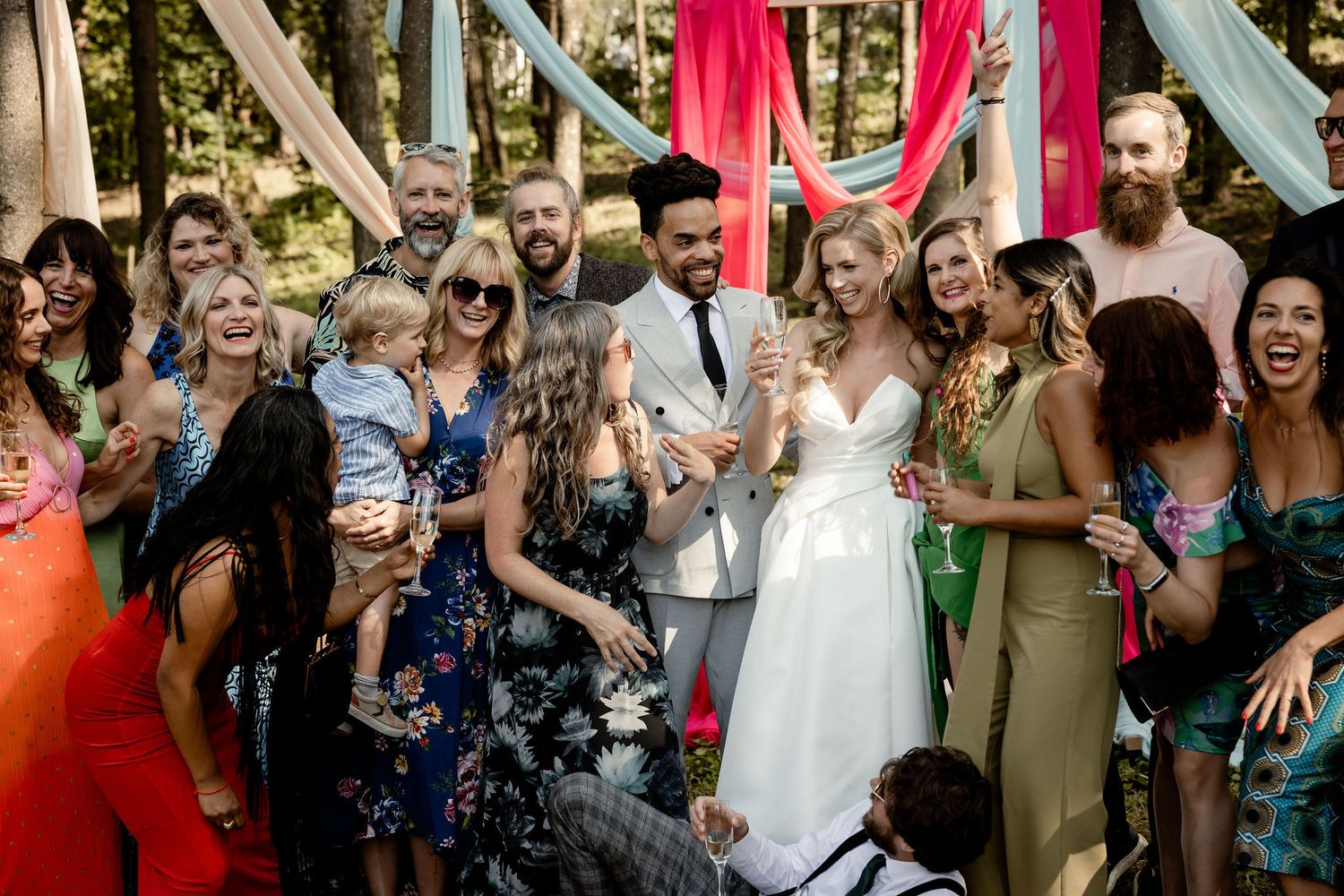 Wedding party celebrates with confetti in a sunlit forest setting with decorative fabric backdrops.