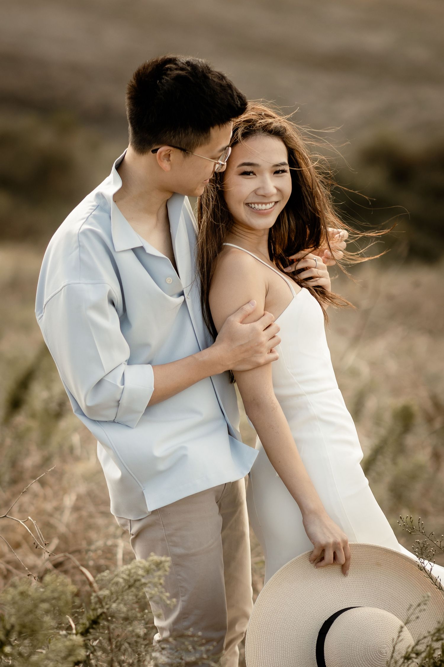 A romantic couple in coordinated white outfits embracing on a wind-swept hilltop at sunset.