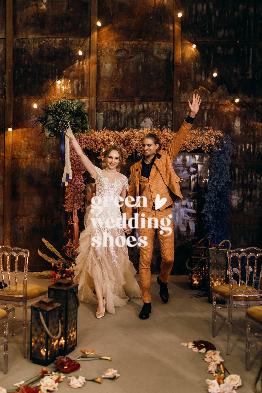 Wedding couple dances joyfully in a rustic venue decorated with warm lighting and floral arrangements on the floor.