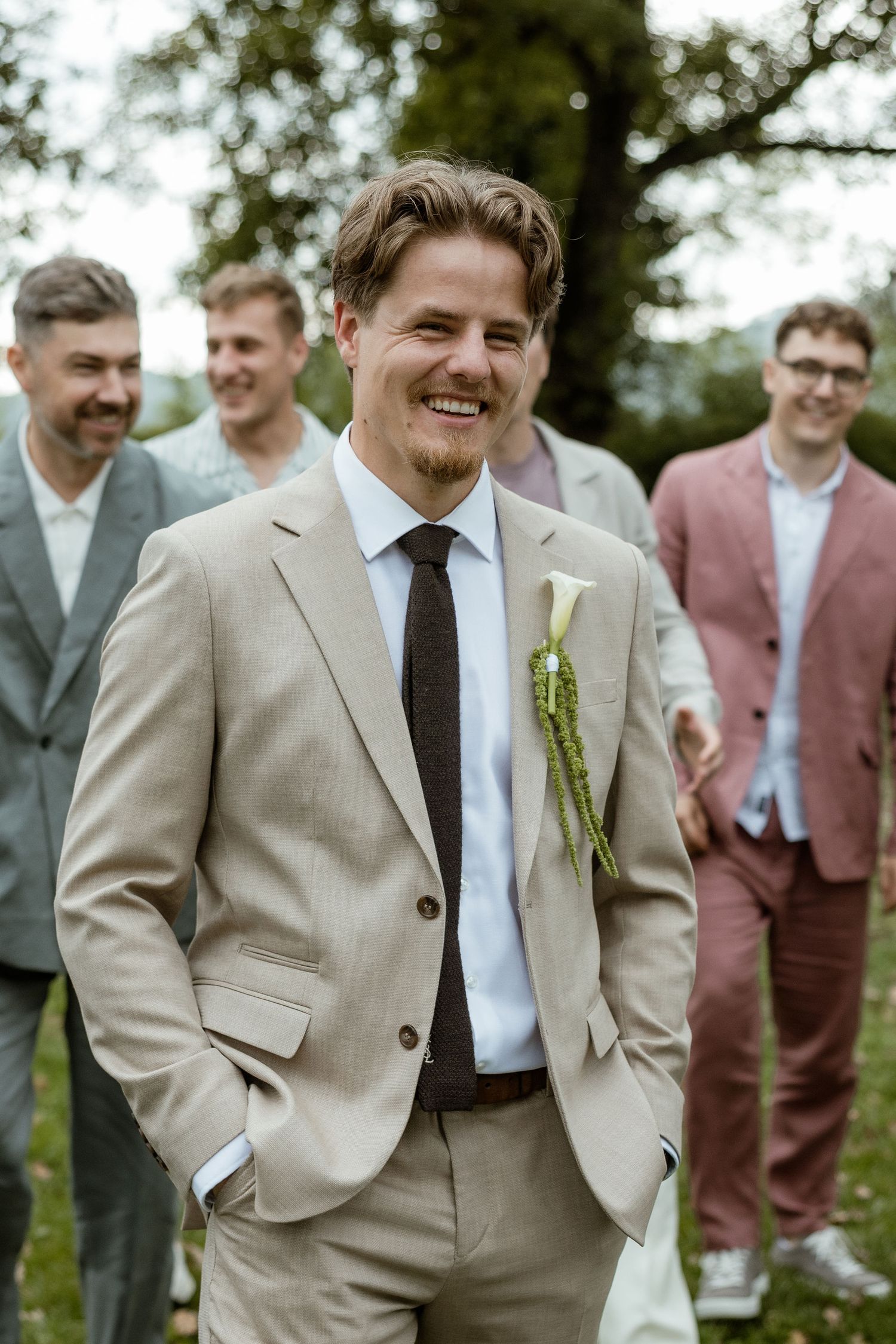 A group of groomsmen in beige suits and ties pose together outdoors during a wedding celebration.