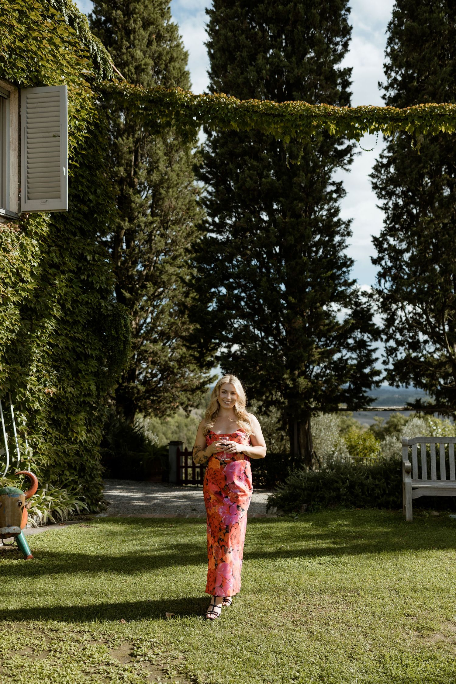 A person in a floral pink dress walks across a sunny lawn with trees and a white building in the background.