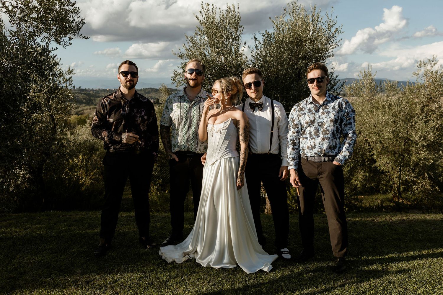A wedding party stands together in a scenic outdoor setting with rolling hills and olive trees in the background.