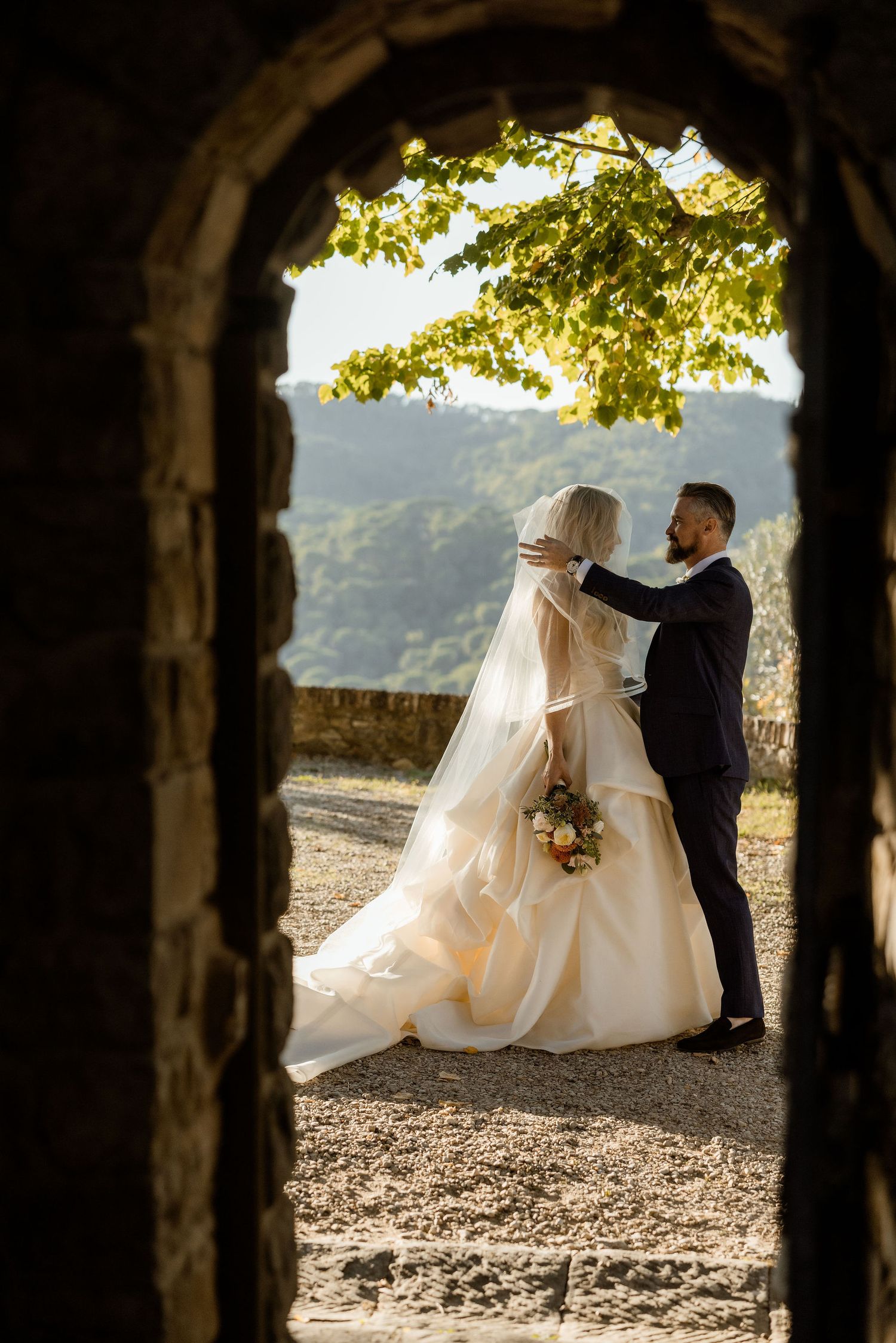 A wedding couple sits on a stone wall framed by trees at sunset creating a romantic silhouette in nature.