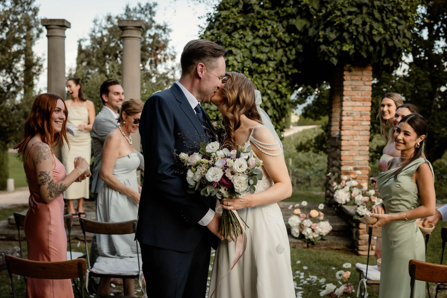 A couple kisses during an intimate outdoor garden wedding ceremony surrounded by smiling guests and stone columns.