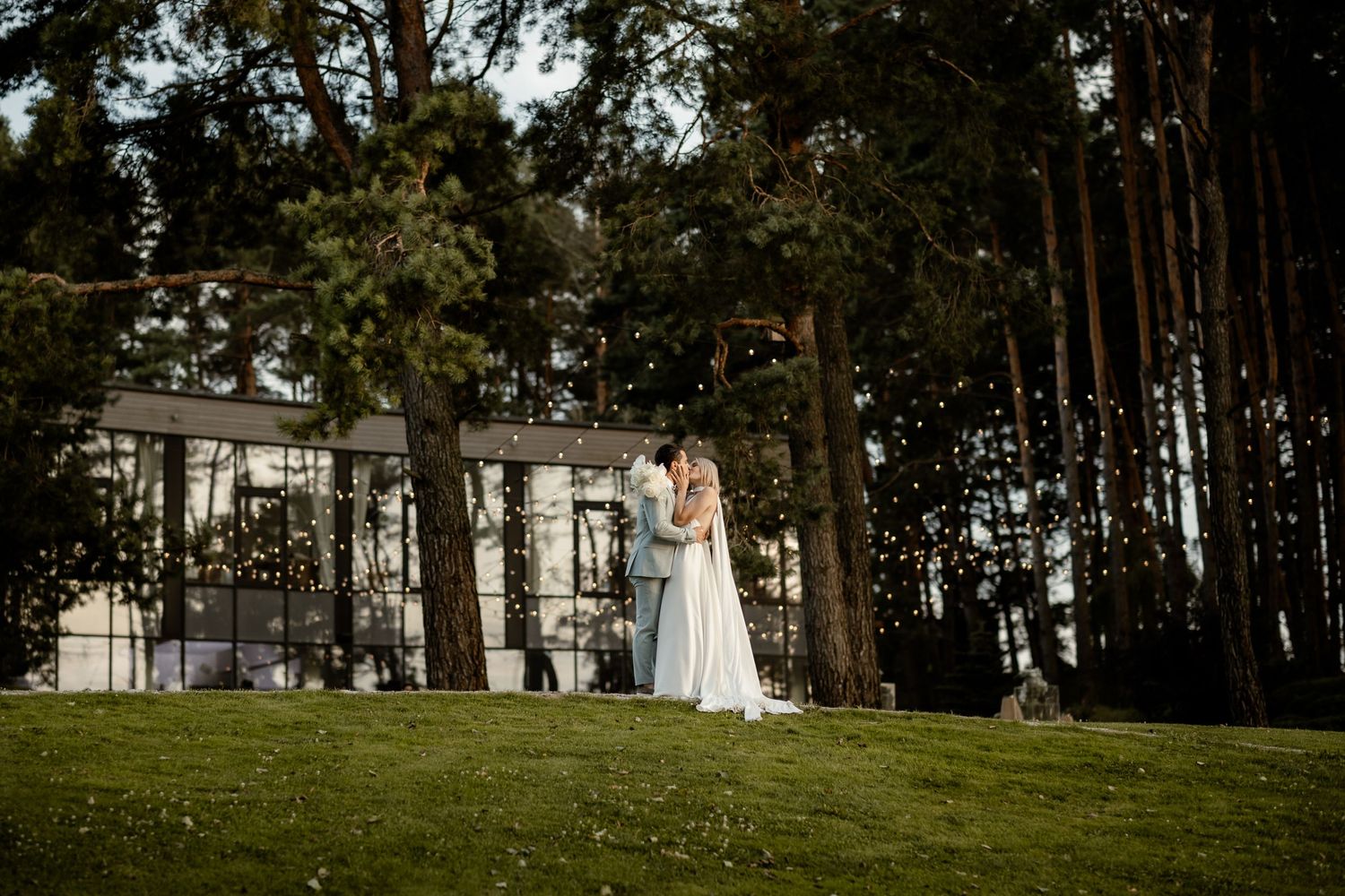 A romantic couple shares a tender moment on a grassy lawn surrounded by tall pine trees near an elegant white mansion.