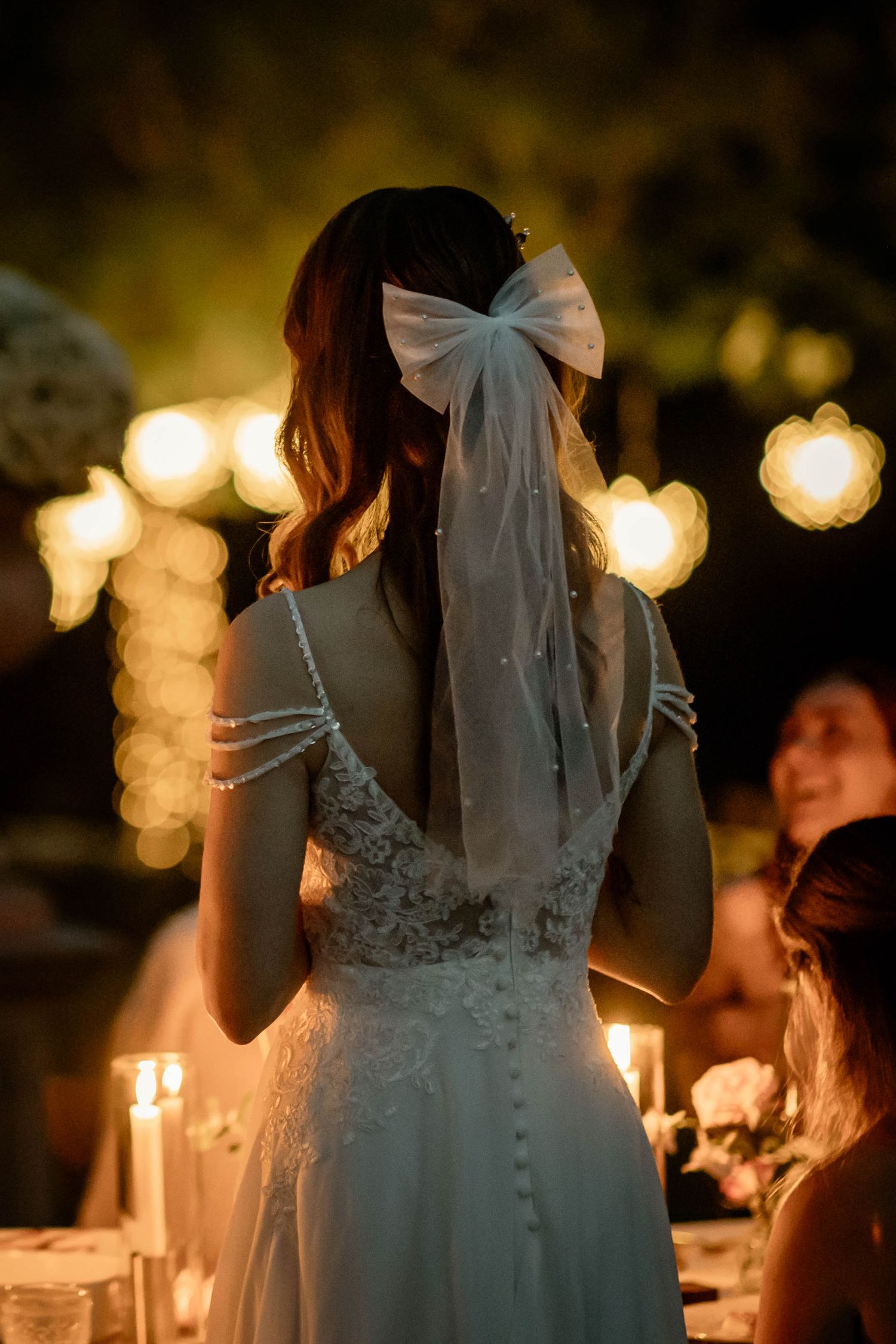 Back view of bride in lace dress with sheer bow headpiece at evening reception with glowing lights in background.