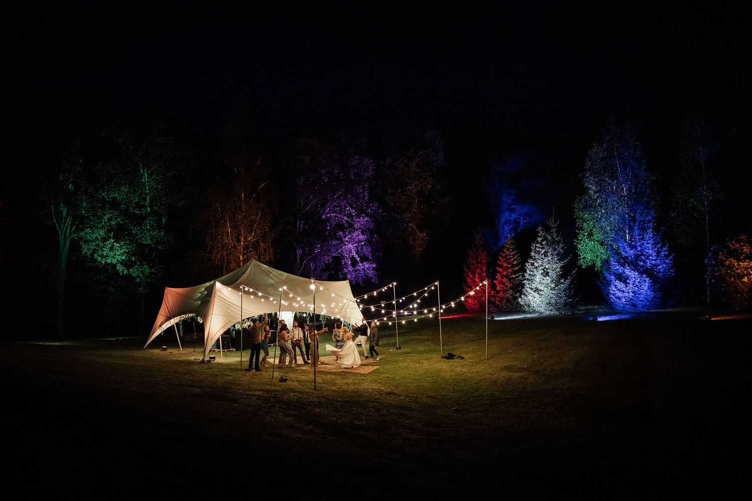 A white tent illuminated at night with colorful lights shining on trees in the background and string lights overhead.