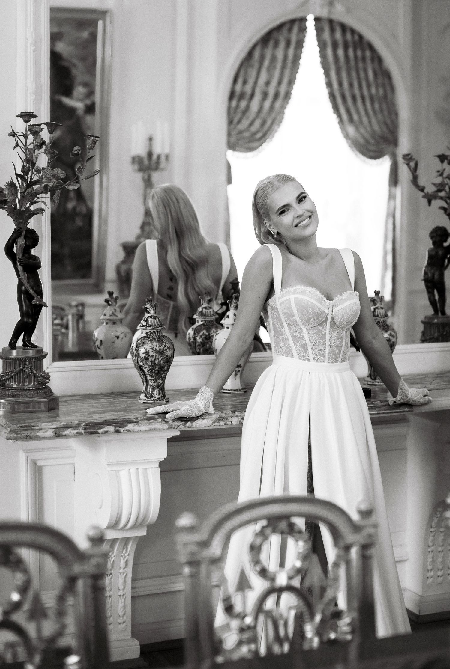 A bride in a structured white gown smiles while leaning against an ornate console table.