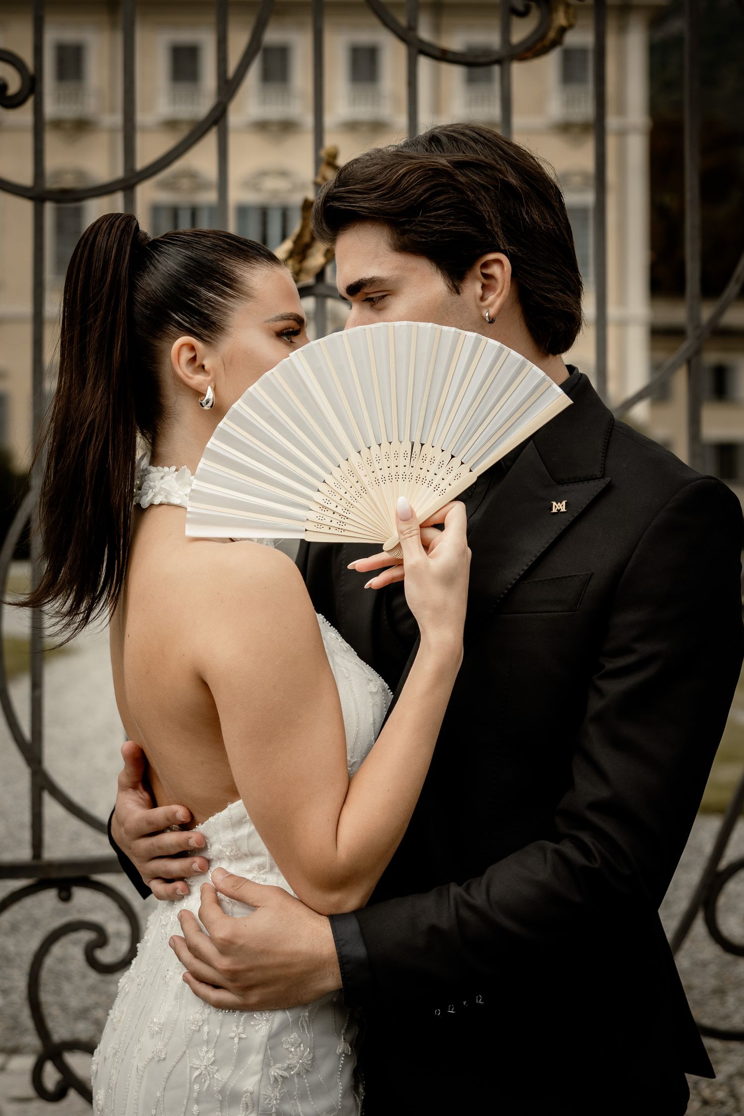 Romantic wedding couple share intimate moment behind white lace fan against ornate iron gates.