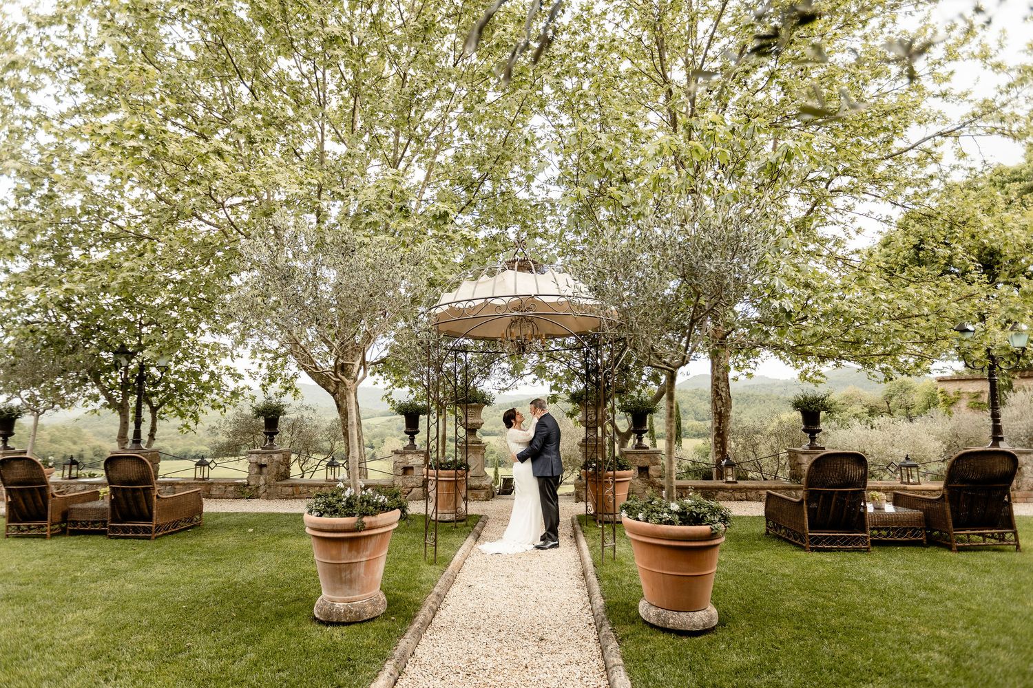 A romantic outdoor wedding ceremony setting with a stone path leading to a gazebo surrounded by potted plants and trees.