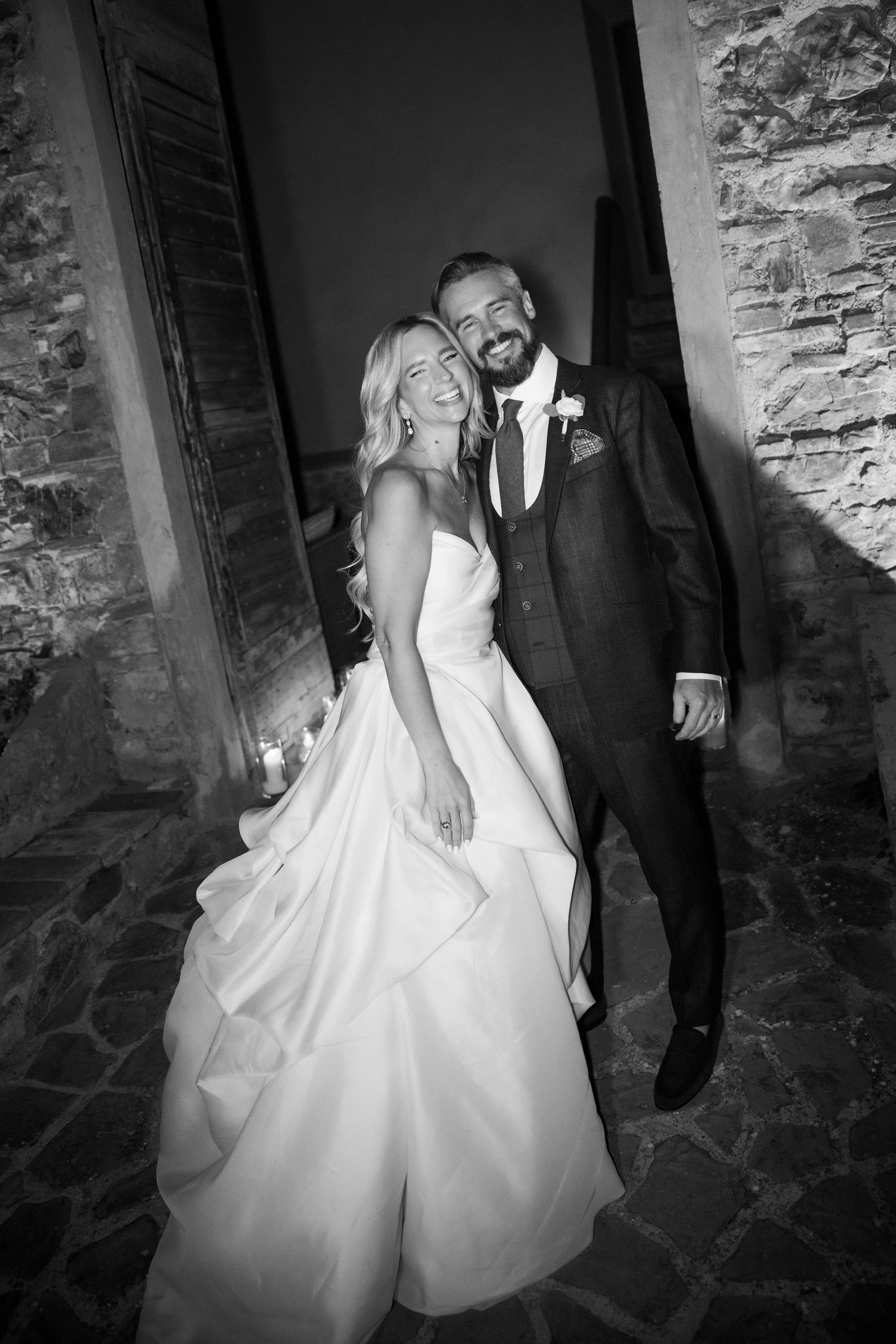 A black and white photo of a couple in wedding attire posing against a rustic stone wall.