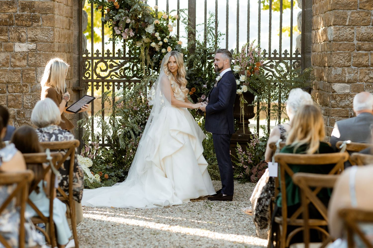 Elegant outdoor wedding ceremony setup with wooden chairs and stone walls framed by decorative iron gates.