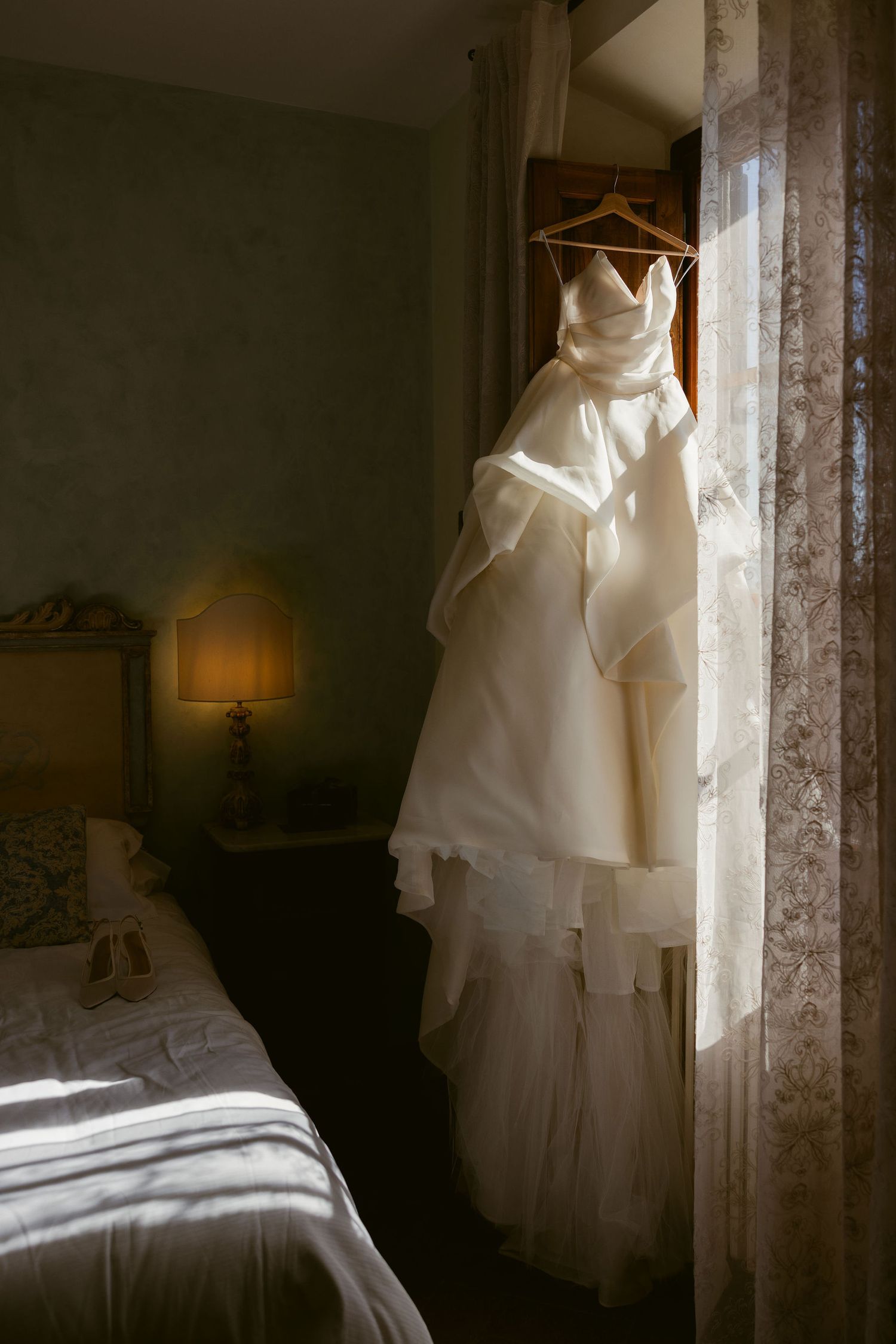 A white wedding dress hangs elegantly in a window, casting soft shadows across a serene bedroom interior.