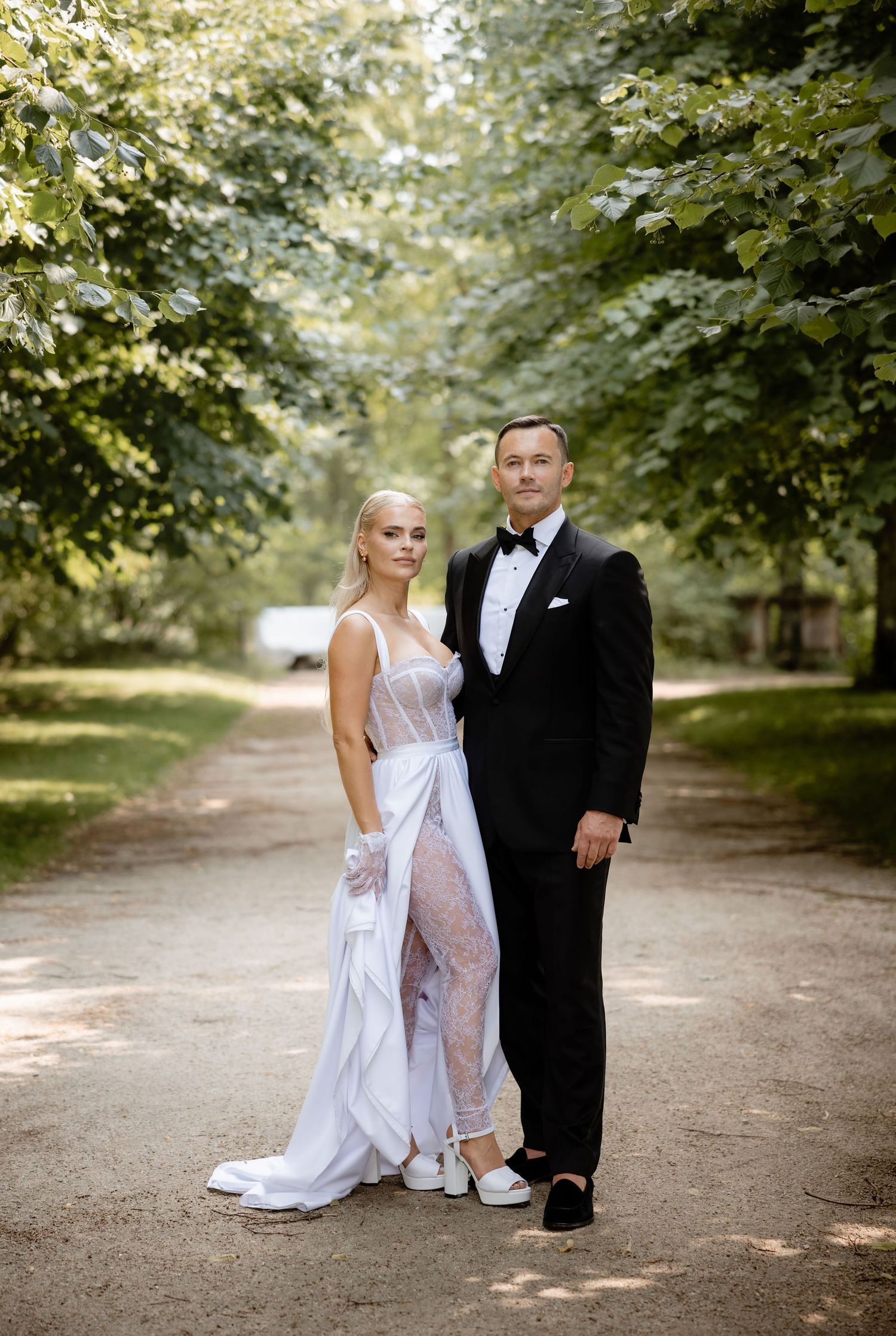 Wedding couple poses on a tree-lined path in formal attire.