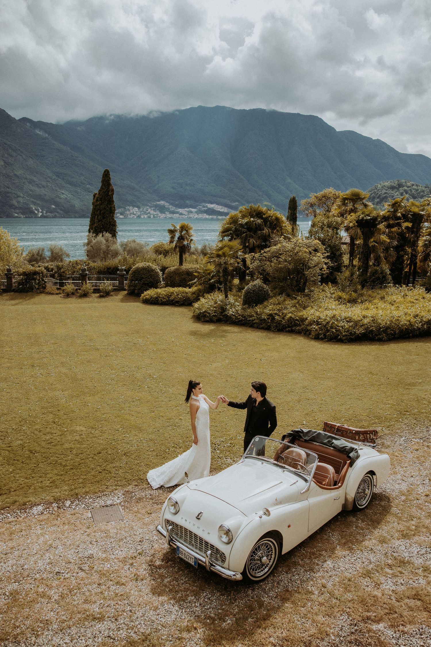 Wedding couple poses with vintage white Triumph car against mountainous lake backdrop.