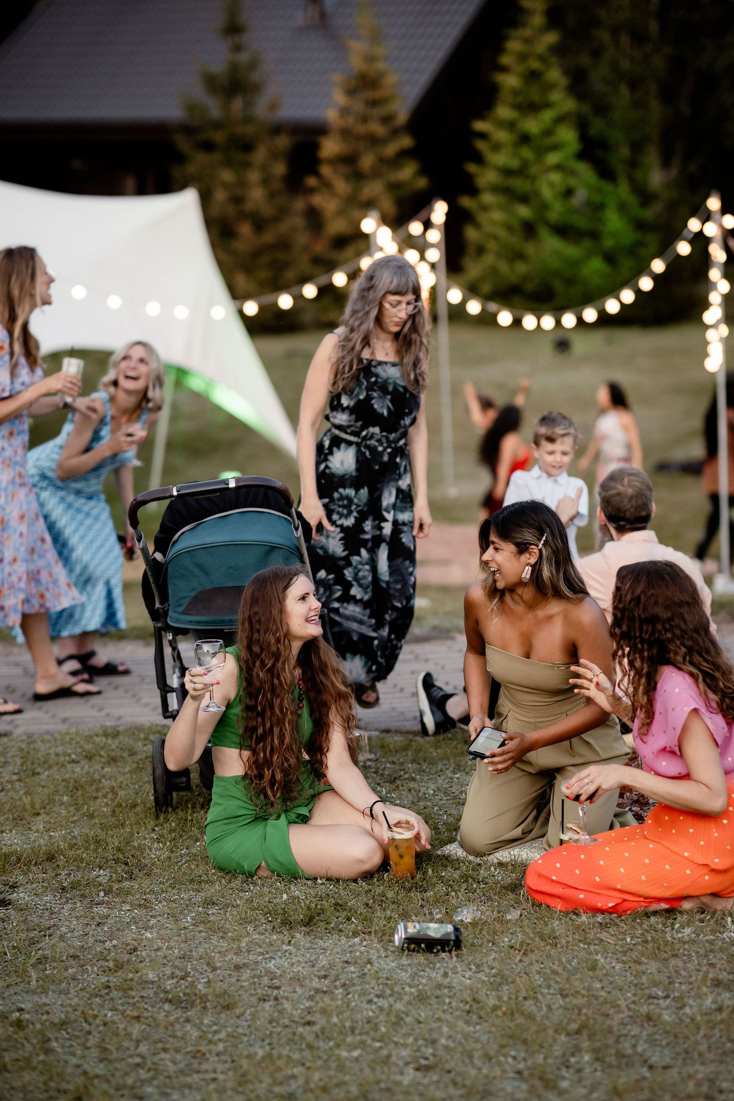 A group of guests enjoying an outdoor summer party with string lights and evergreen trees in the background.