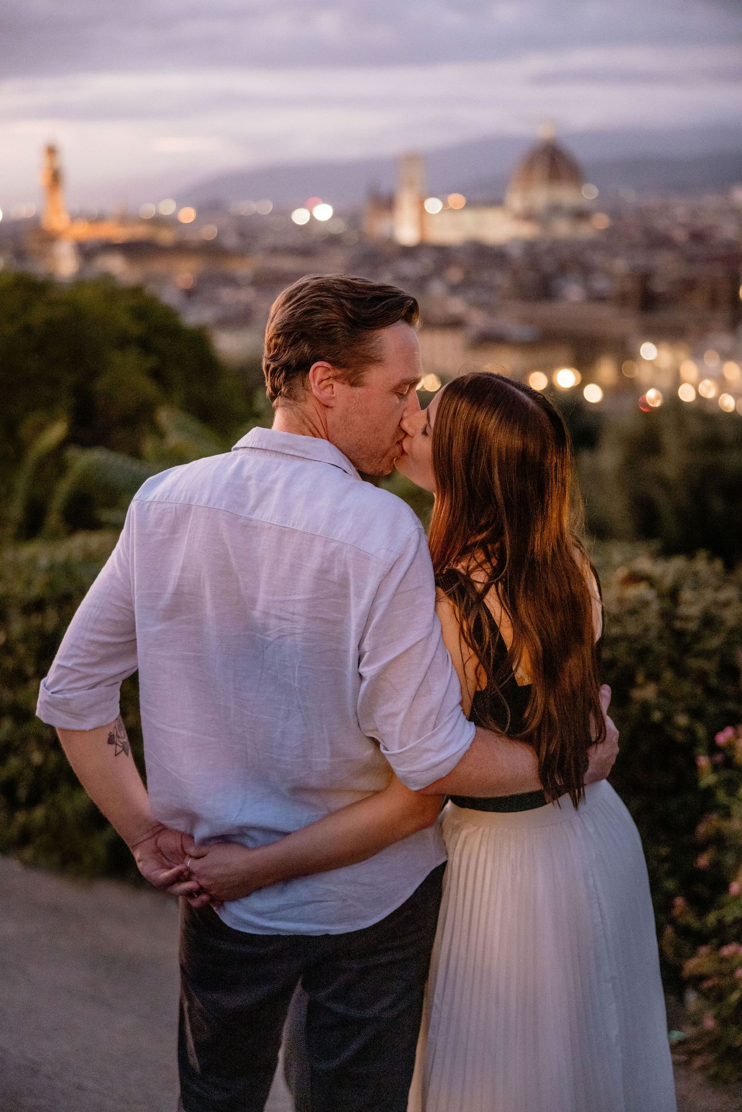 A romantic couple sharing a kiss at sunset overlooking the Florence skyline with the Duomo cathedral visible in the distance.