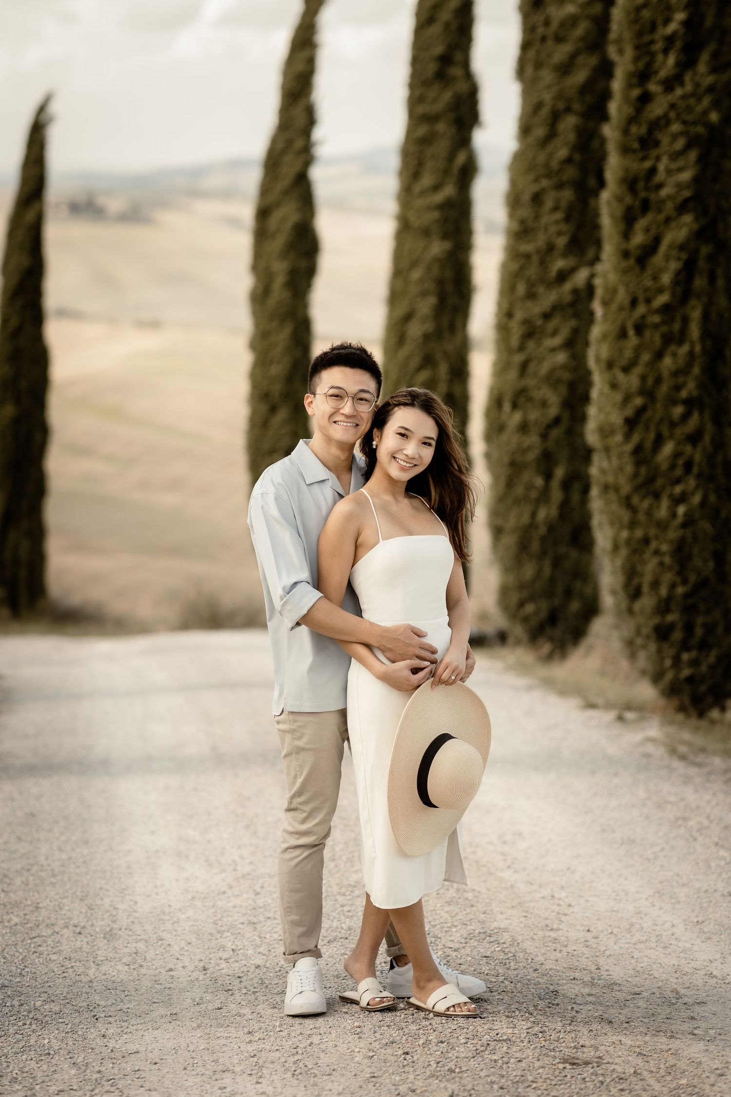 Stylish couple in coordinated outfits pose on scenic Tuscan cypress tree road.