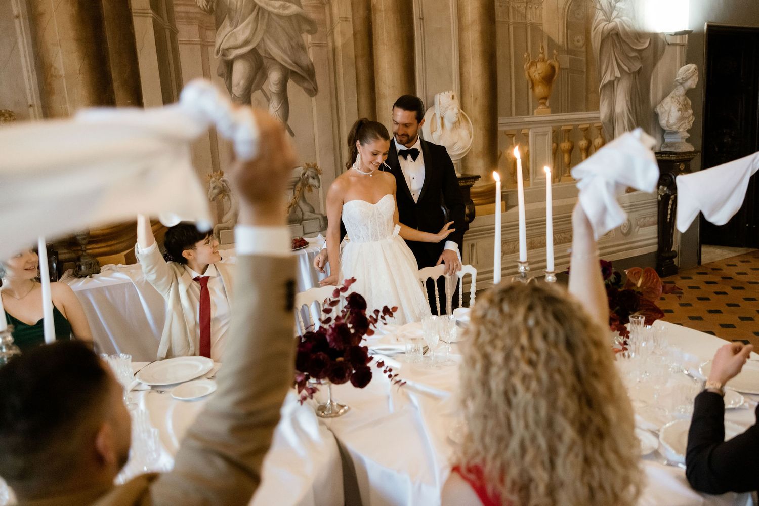 Elegant wedding reception in a vintage hall with white decor and tall candles while guests raise toasts at tables.