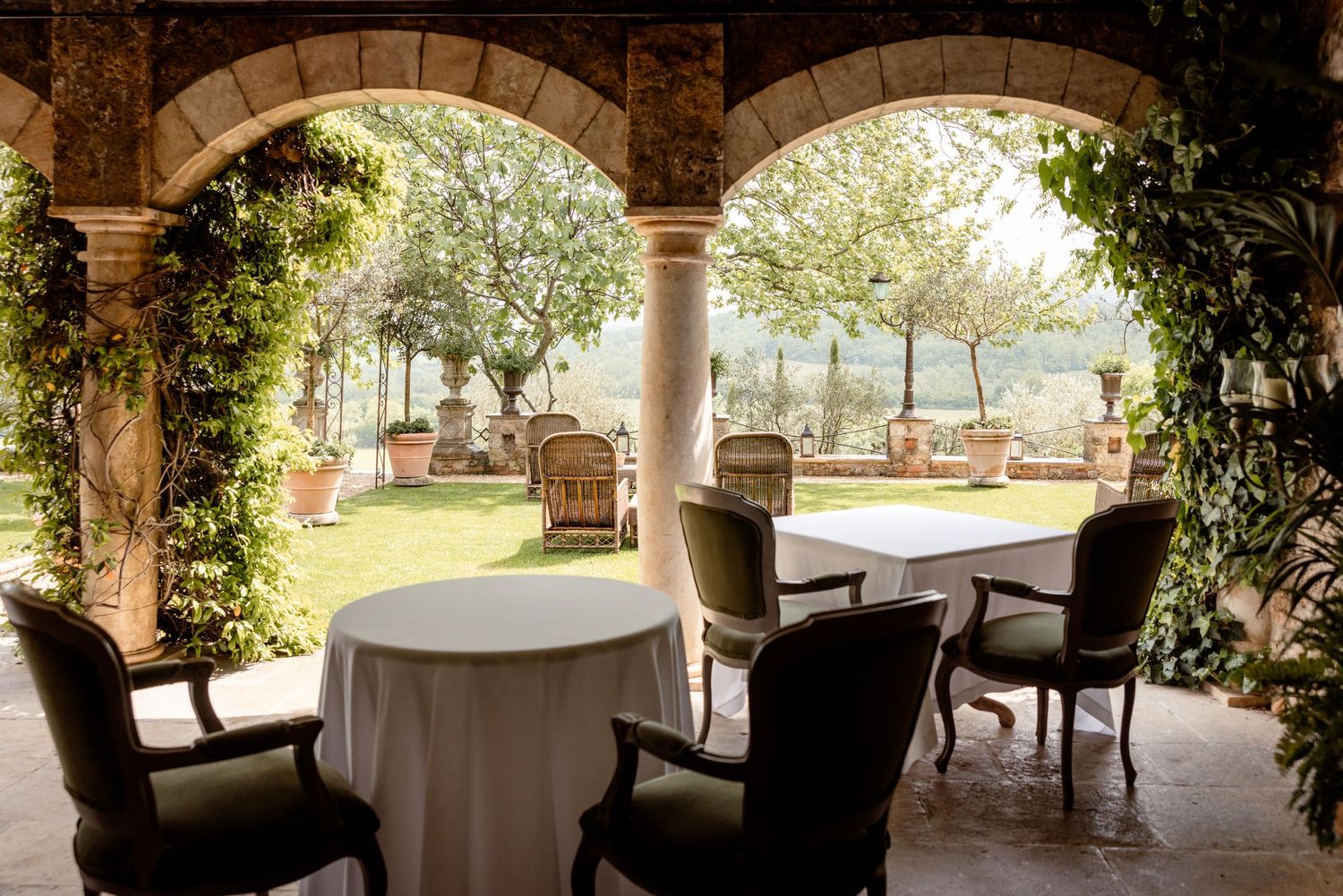 Elegant outdoor dining terrace with stone columns, white tablecloths and wicker chairs overlooking a sunny garden patio.