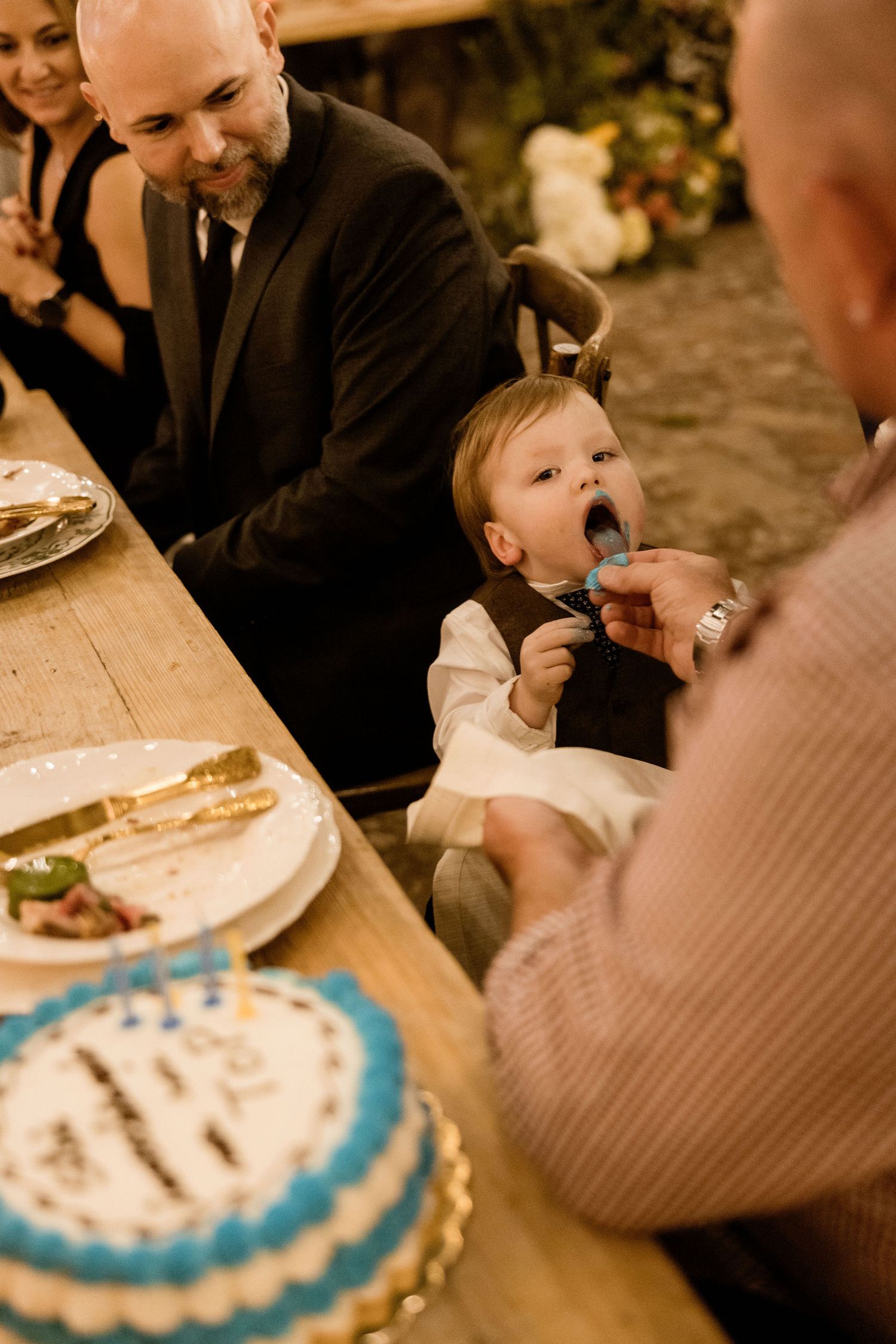 Guests gathered around a table at a celebration with a blue and white decorated cake in warm candlelight.