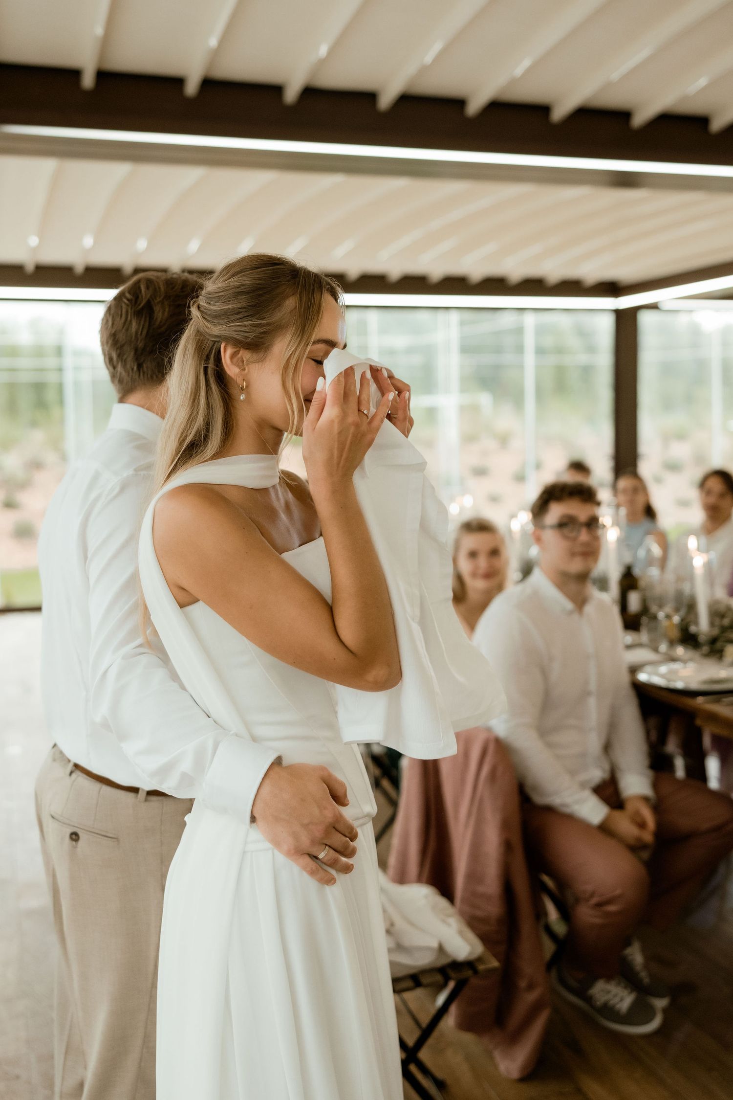 A couple shares an intimate embrace on their wedding day while guests look on in a bright indoor venue.