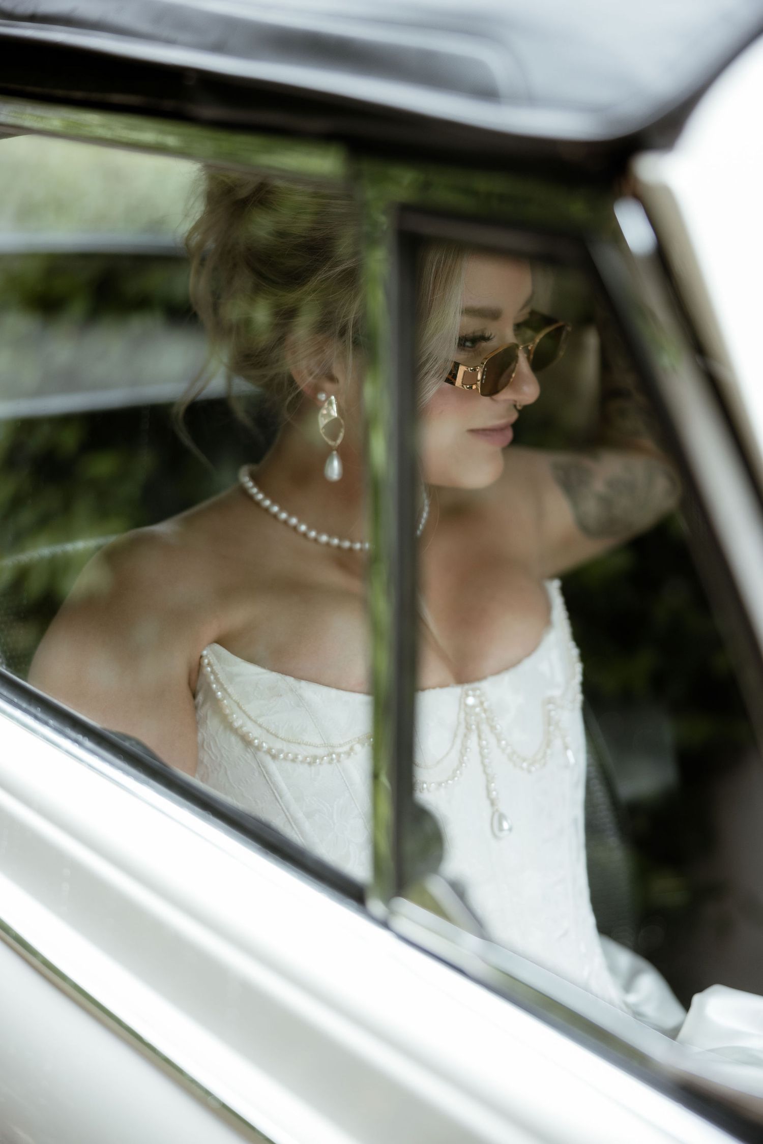 Through a car window, a bride in a white dress with pearl necklace and earrings sits elegantly in vintage style.