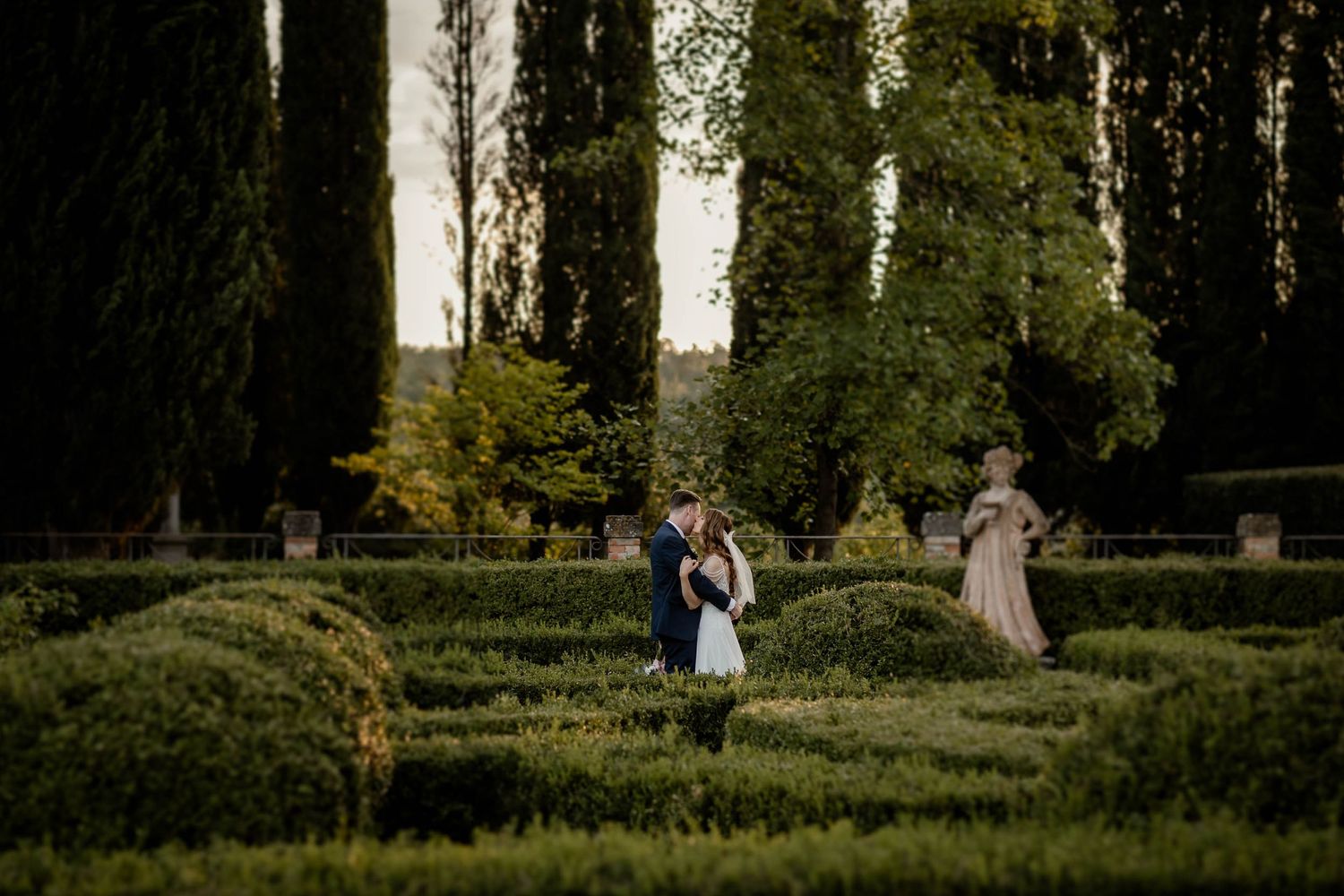 Newlyweds celebrate in a lush garden setting with tall trees and manicured hedges at sunset.