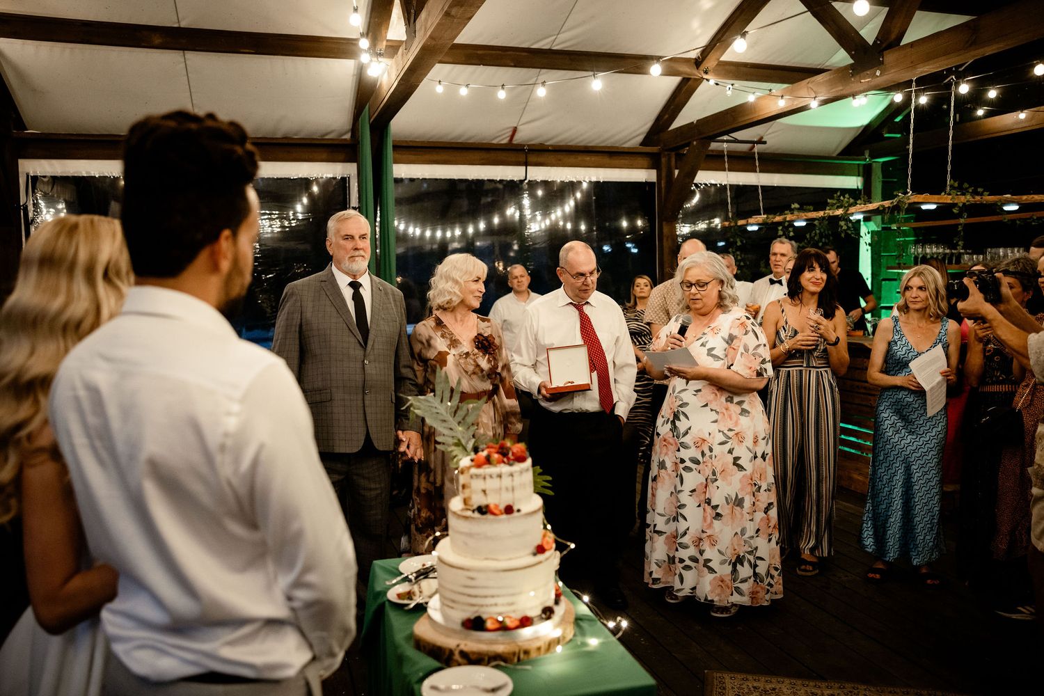 Wedding reception guests gather around a three-tiered cake decorated with flowers in a rustic indoor venue.