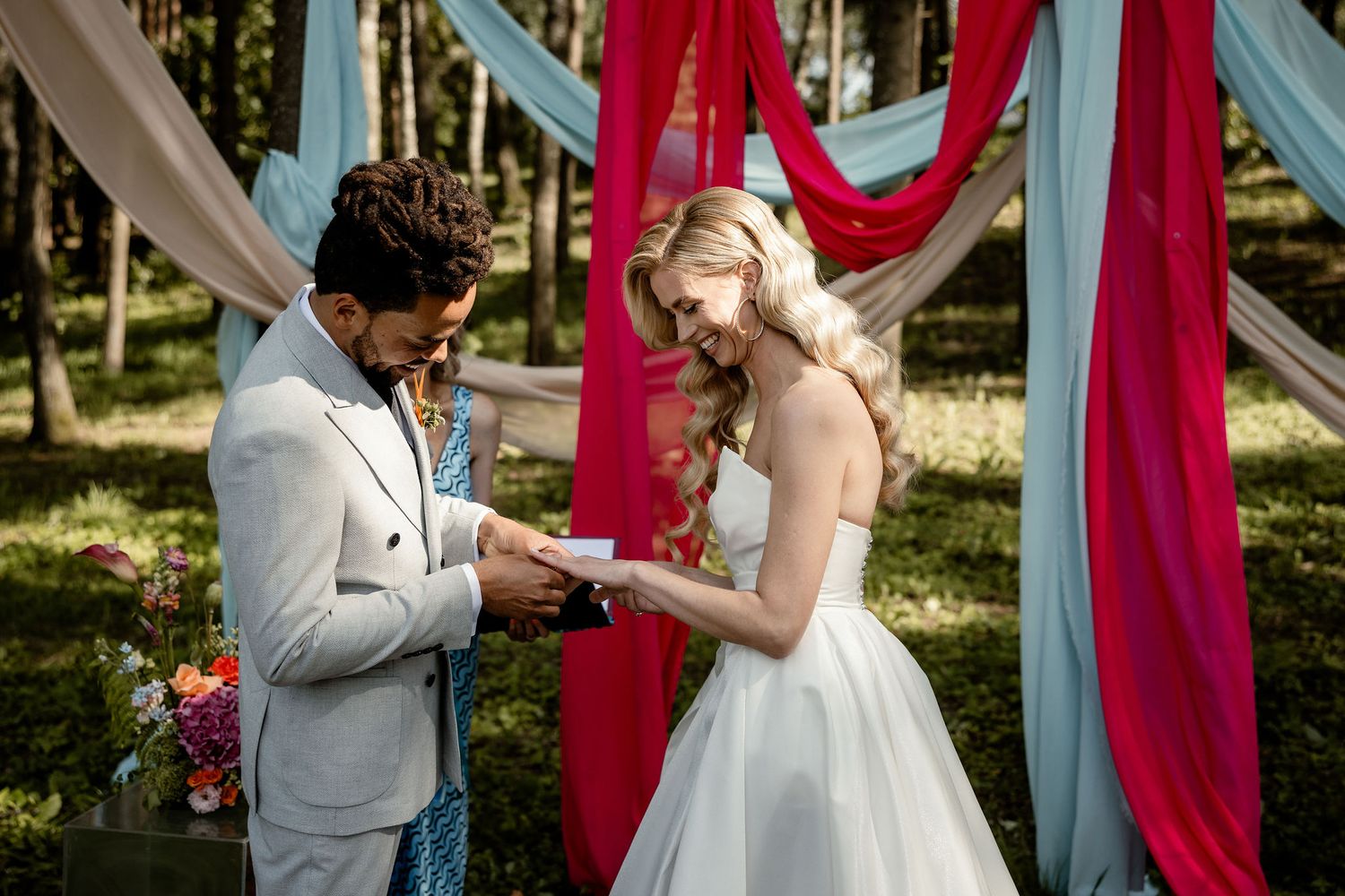 A couple exchanges rings during their outdoor forest wedding ceremony under red and blue draped fabric.