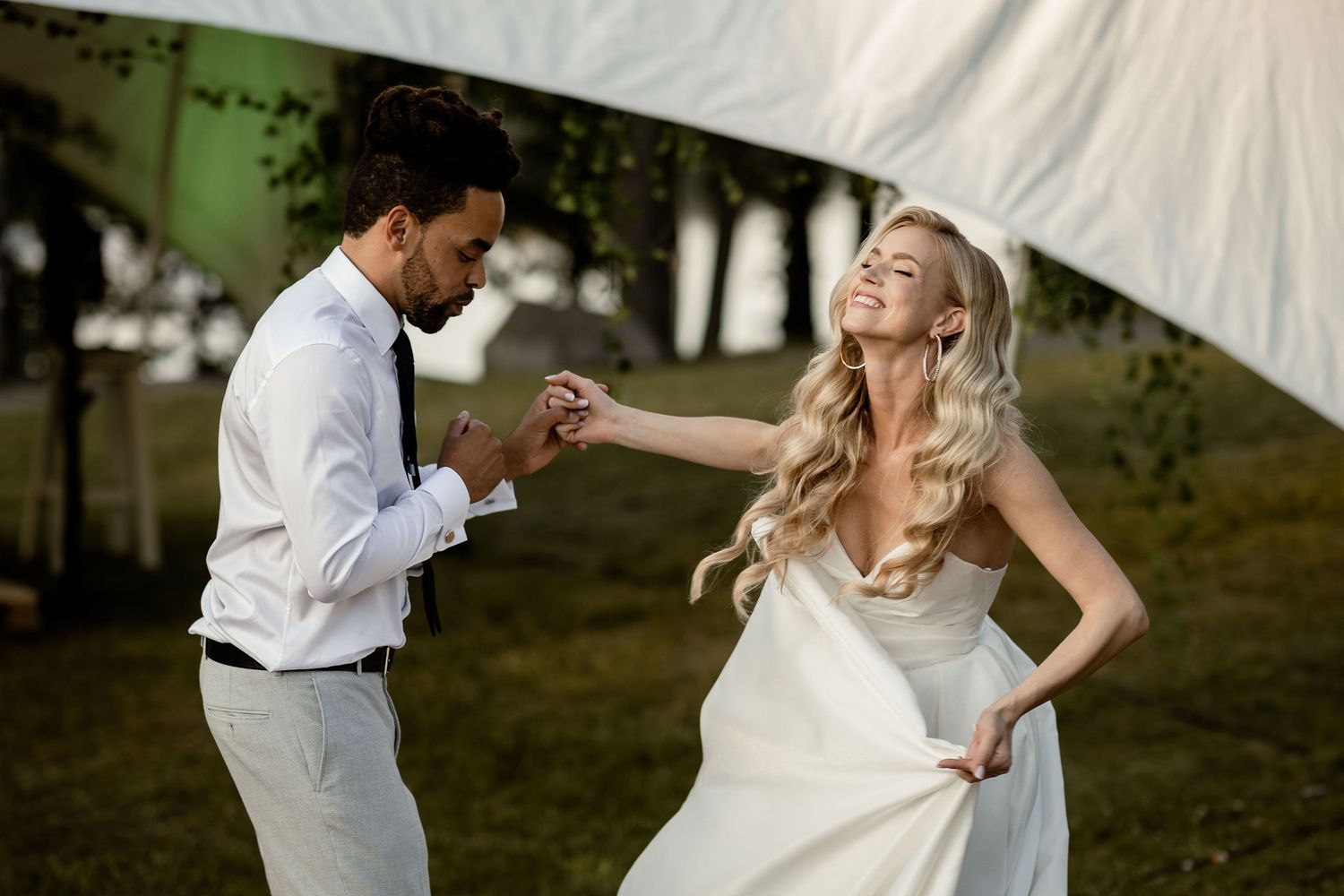 A joyful couple dancing together outdoors under a white tent, the bride wearing a flowing white gown.