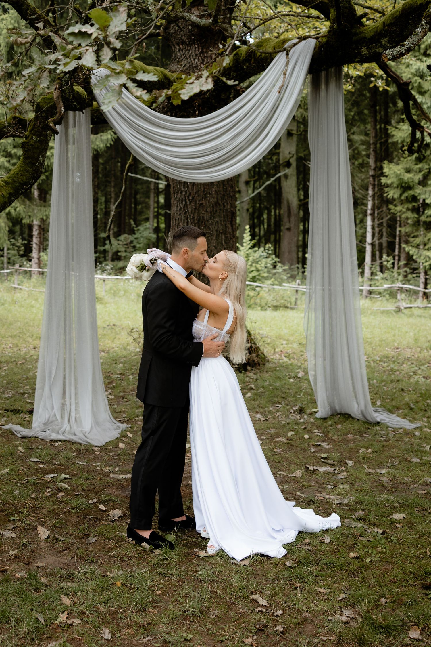 A wedding kiss under an elegant white fabric arch in a forest setting.