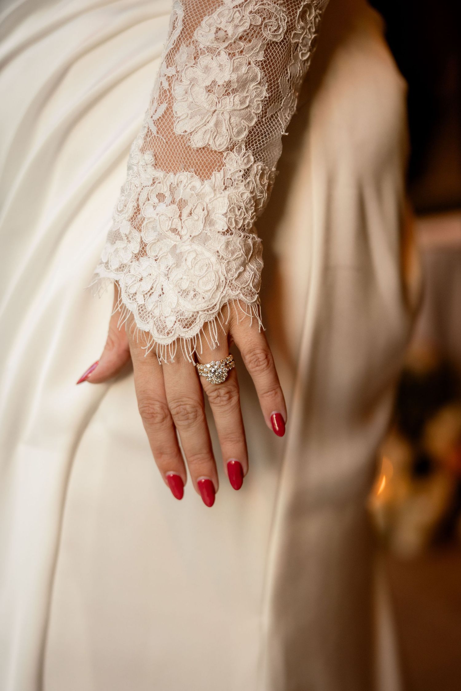 Elegant bridal lace sleeve detail with red manicured nails and diamond engagement ring against white wedding dress.